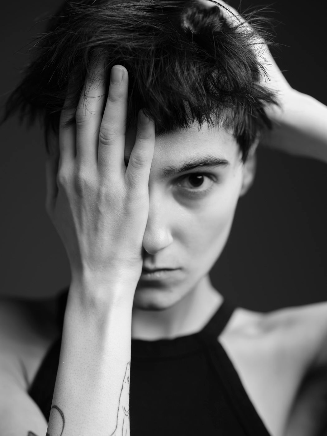 Black and white portrait of a person with short hair covering one eye with a tattooed hand.