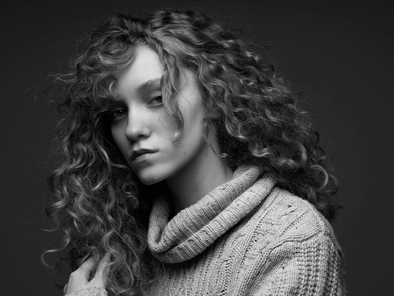 Black and white portrait of a young woman with curly hair wearing a cozy knit turtleneck sweater.