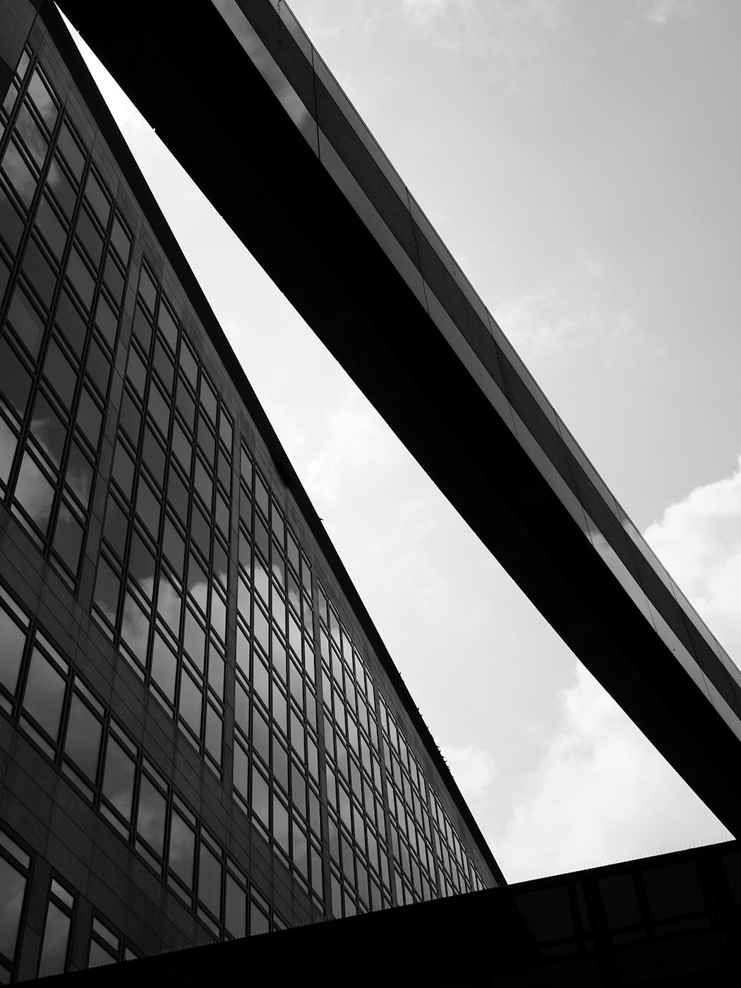 Black and white architectural photo looking up at a curved glass building facade and overpass against a cloudy sky.