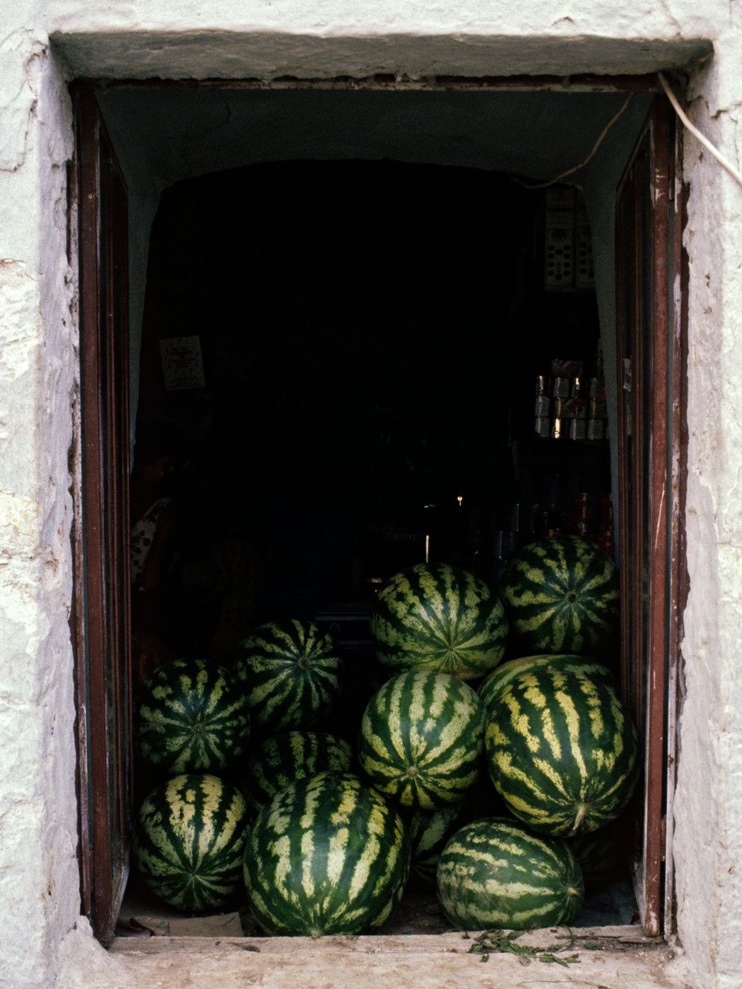 Pile of fresh watermelons displayed in a rustic white stone doorway of a small market shop.