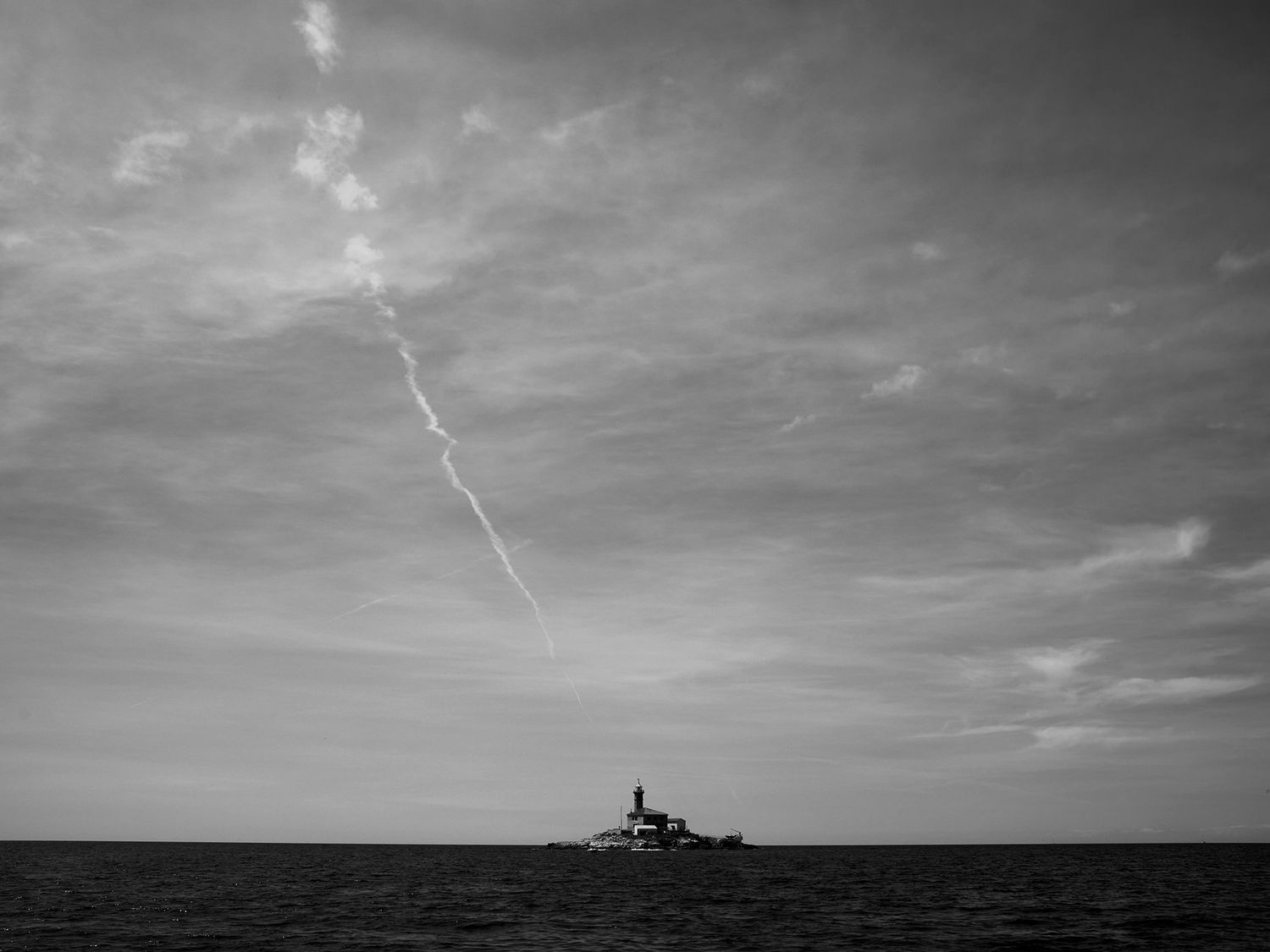 Black and white photo of a lone lighthouse on a small island in a calm sea under a dramatic cloudy sky.