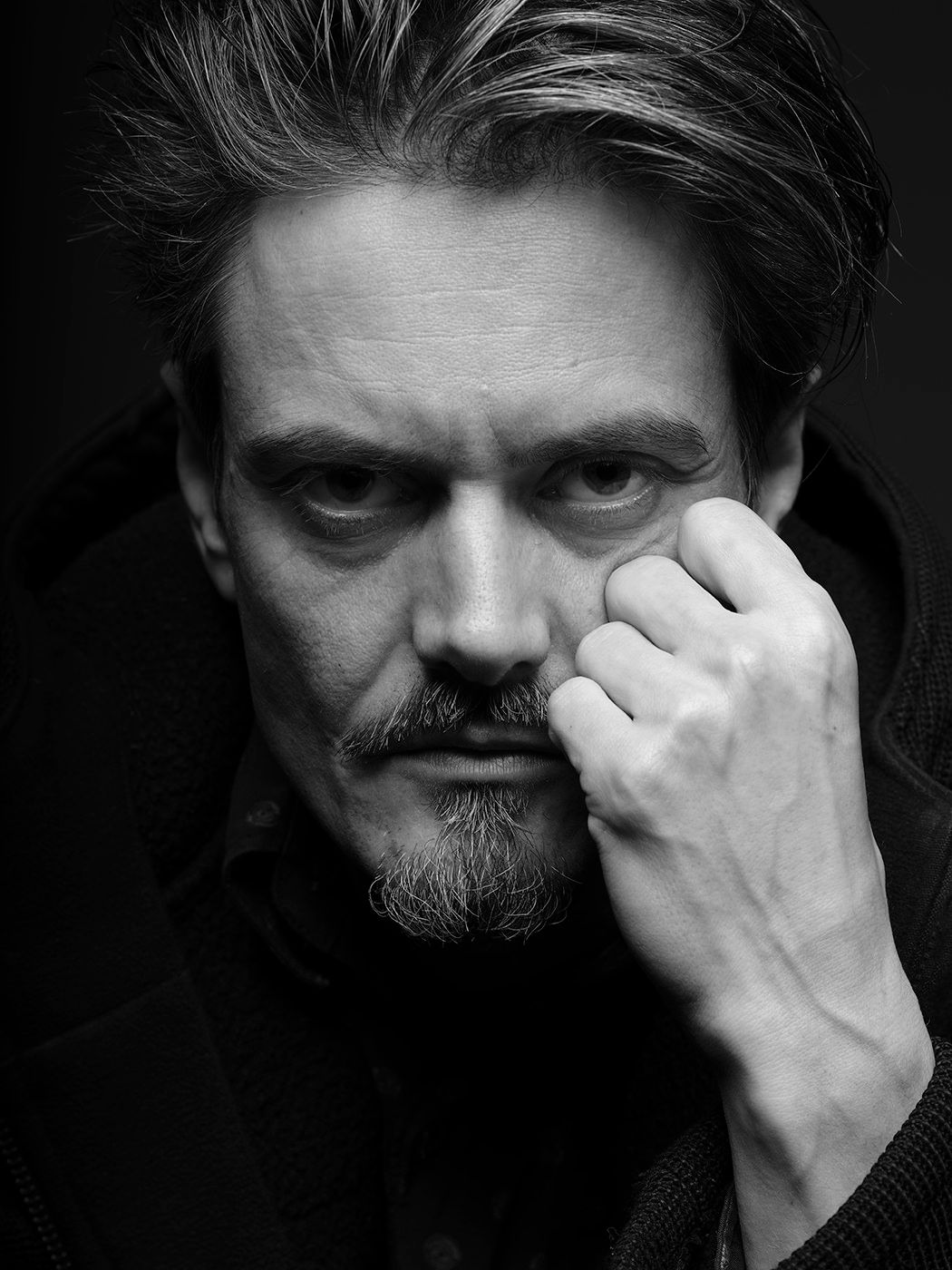 Dramatic black and white portrait of a middle-aged man with slicked hair resting his fist against his chin.