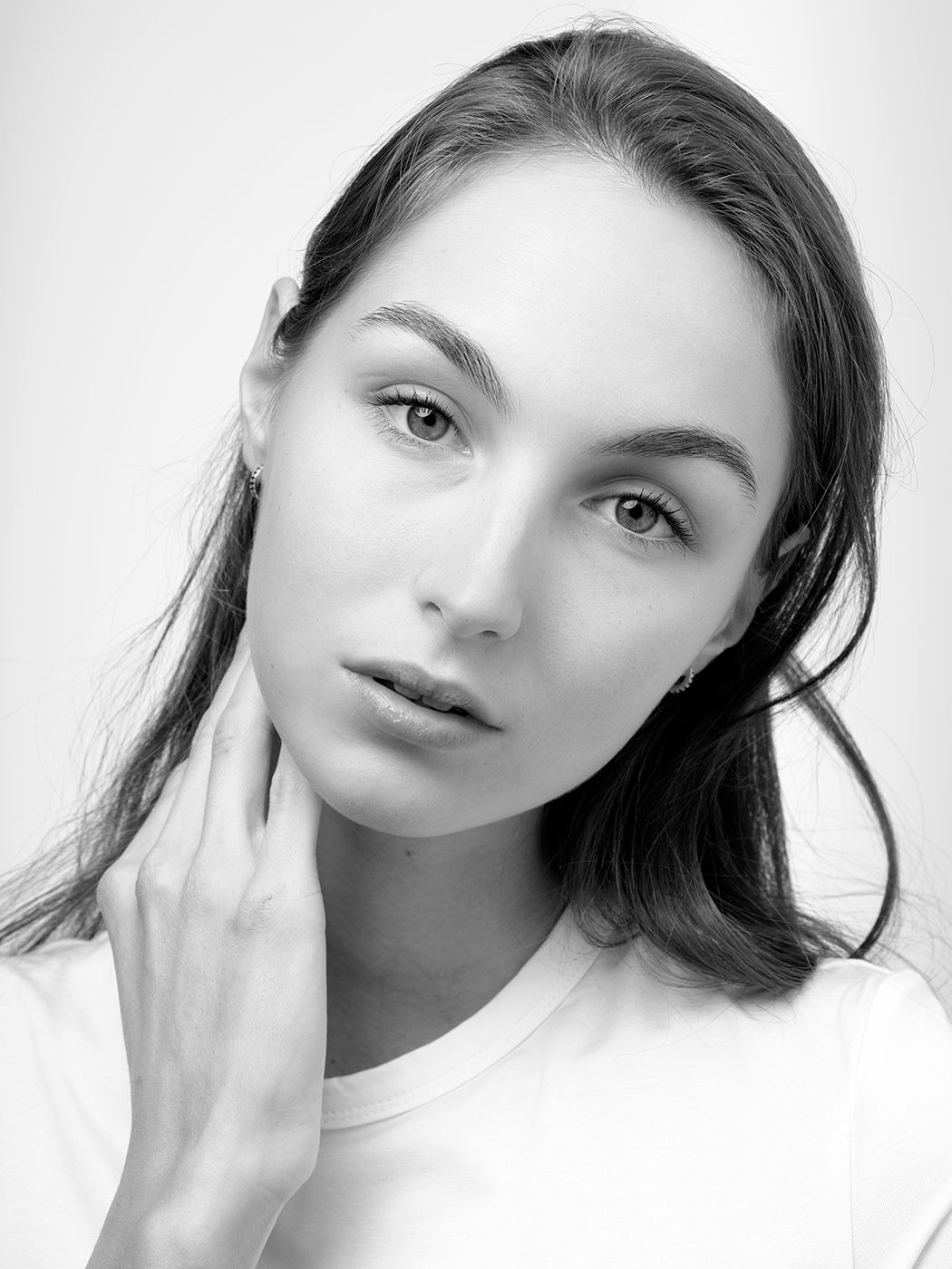 Black and white portrait of a young woman with long dark hair touching her neck, wearing a white t-shirt.