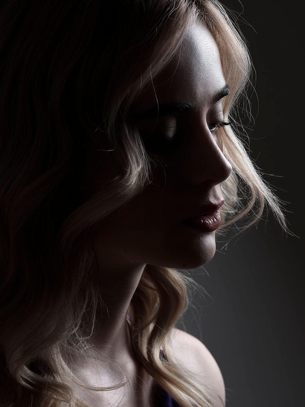 Dramatic low-key portrait of a blonde woman with closed eyes, side profile in moody dark studio lighting.