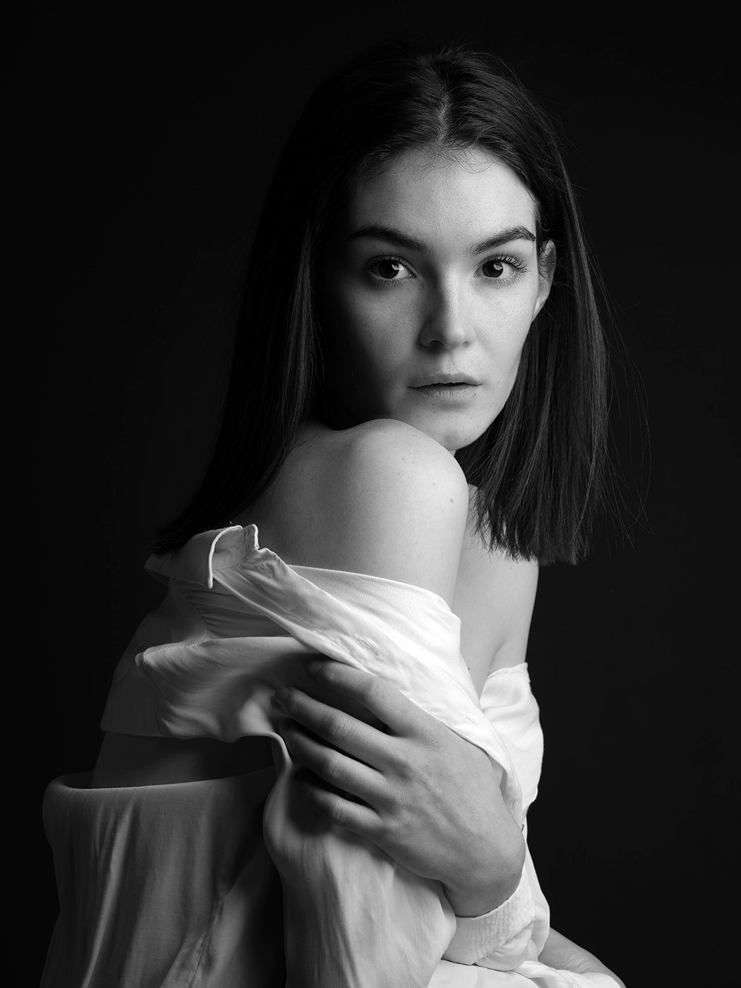 Black and white portrait of a young woman with dark hair, wearing an off-shoulder white shirt, against a dark background.