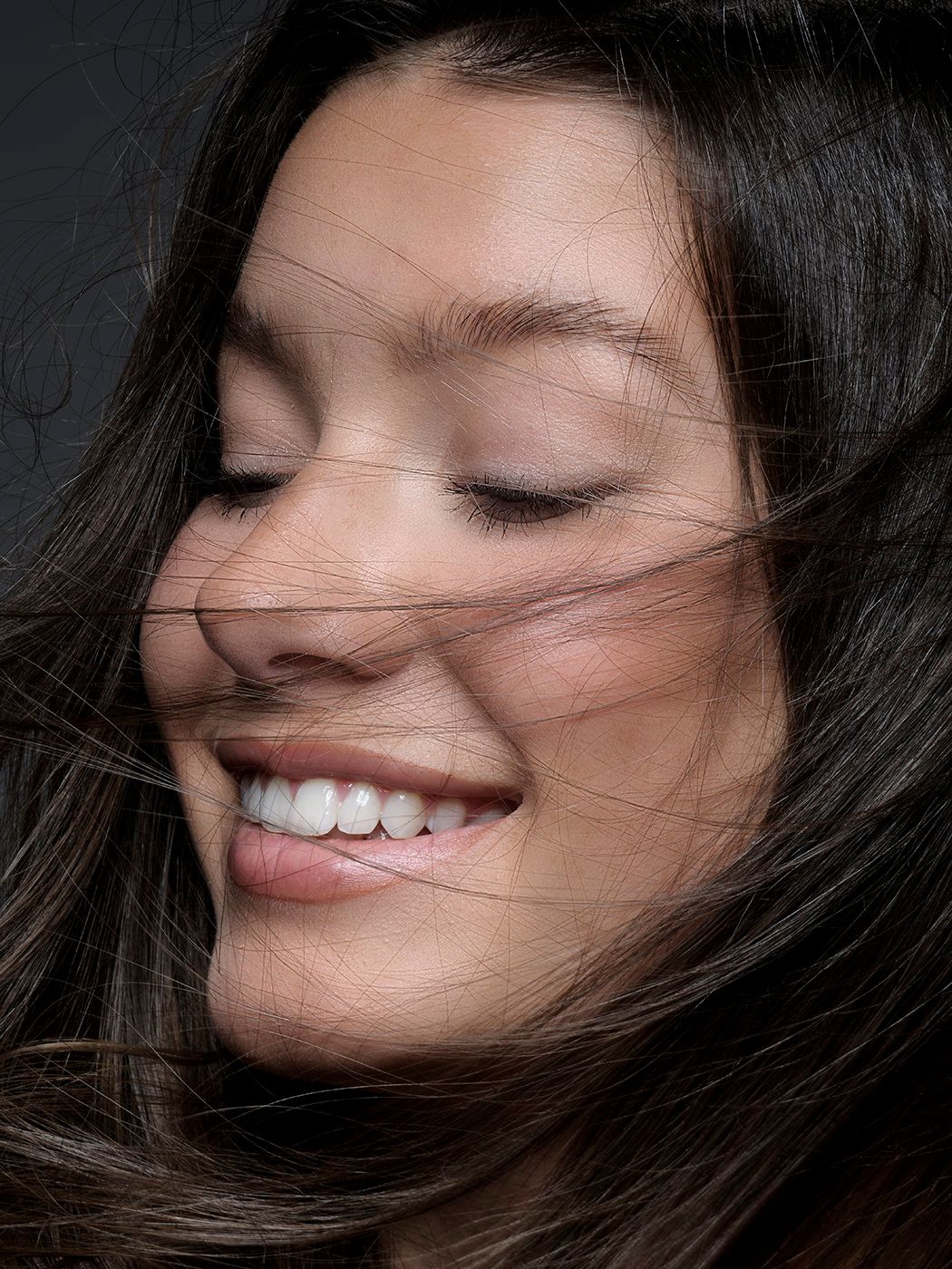 Young woman with long dark hair blowing across her face, smiling with eyes closed against dark background.