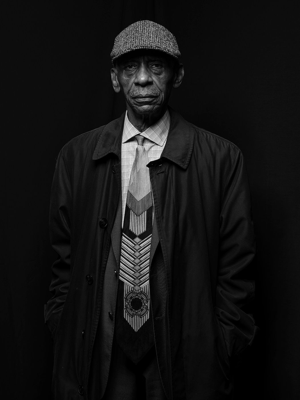 Black and white portrait of an elderly man wearing a flat cap, trench coat, and decorative beaded tie. Roscoe Mitchell, musician, sax