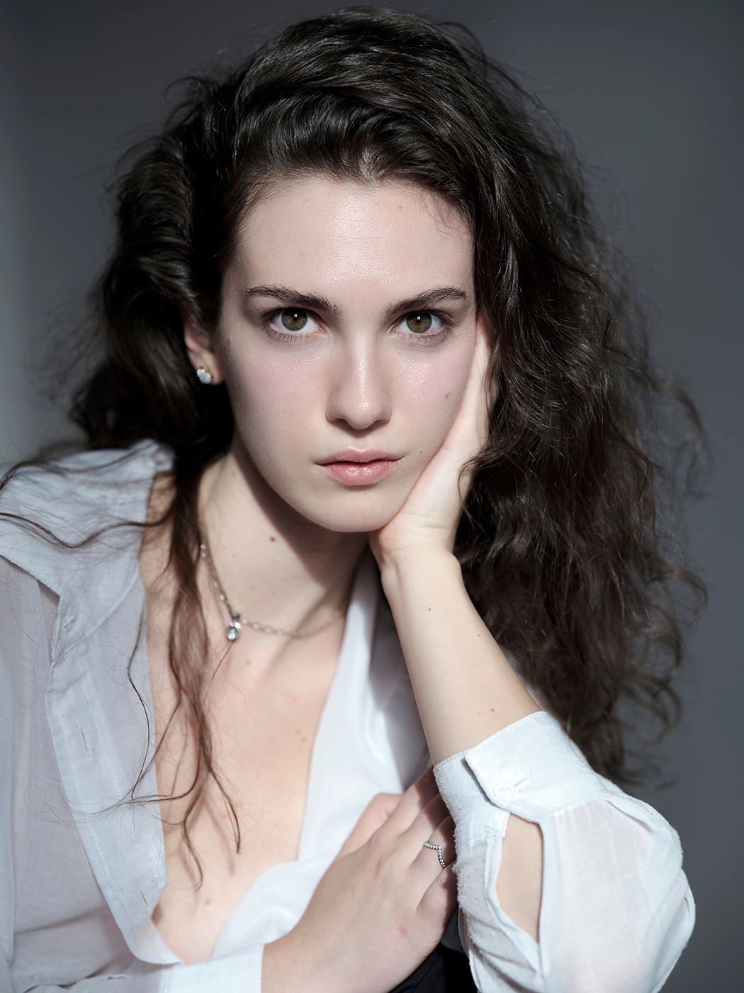 Young woman with curly dark hair wearing a white blouse and necklace, posing with hand against cheek.