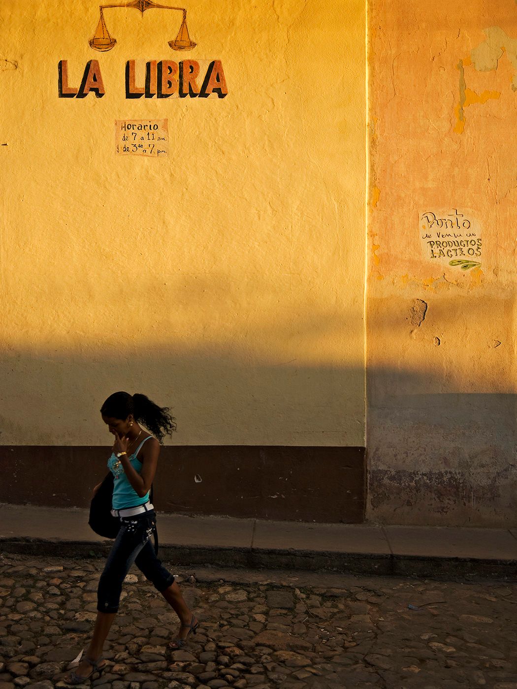 Woman walking past a golden yellow wall with La Libra sign on a cobblestone street.