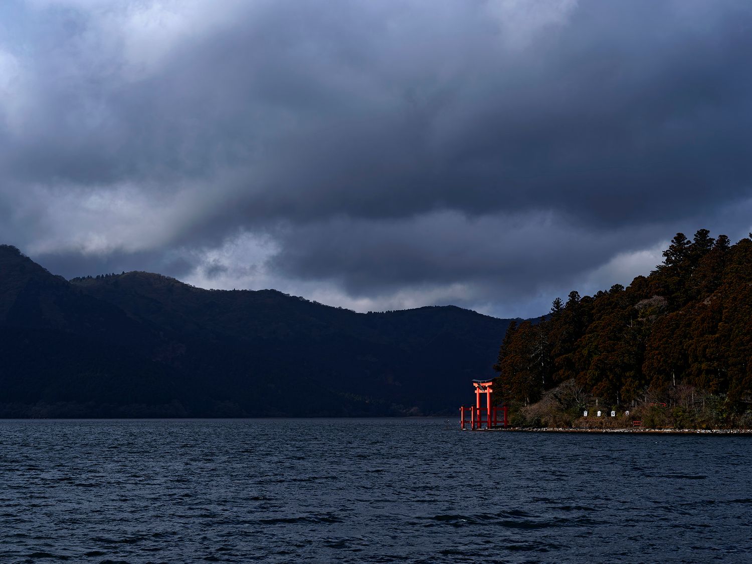 A red torii gate stands in Lake Ashi under dramatic storm clouds with misty mountains in Hakone, Japan.