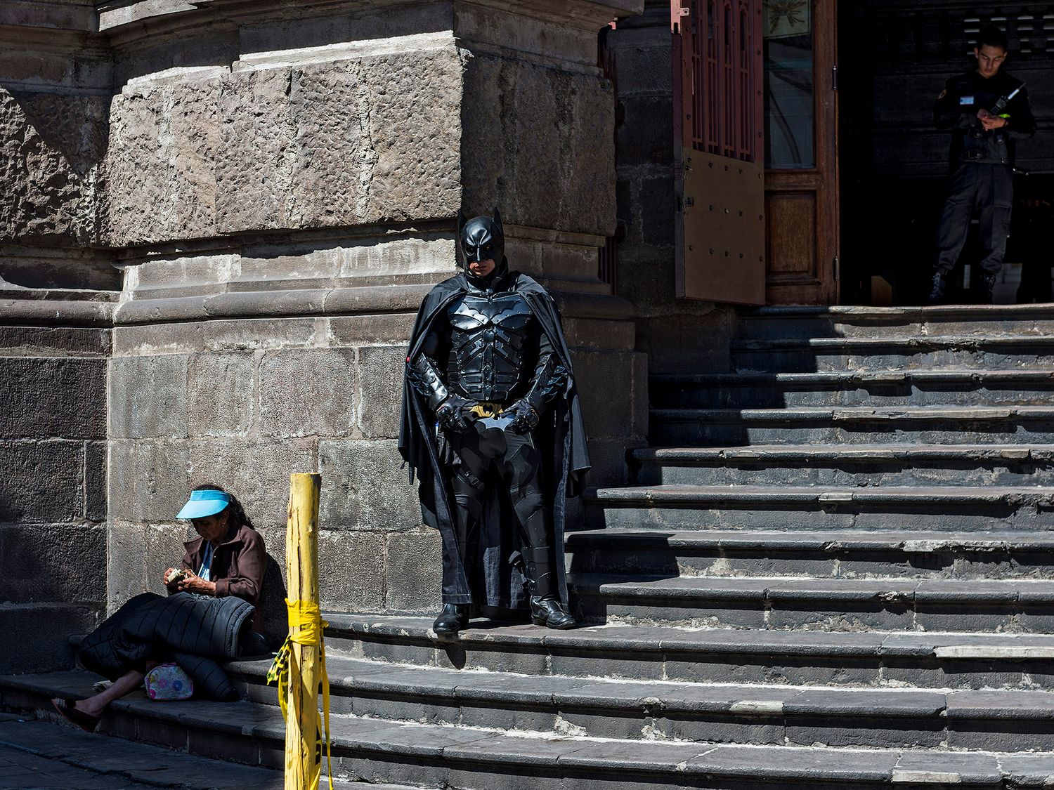 Person in Batman costume stands on stone steps beside a seated woman outside a historic building in bright sunlight.