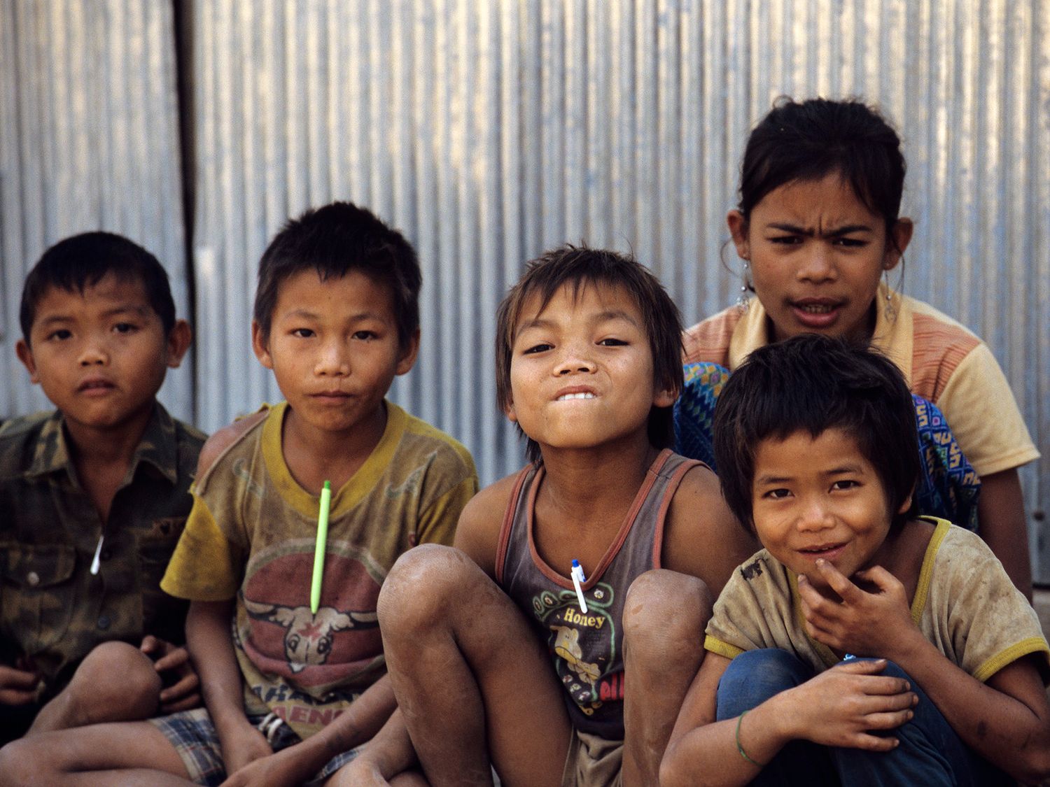 Five young Southeast Asian children sitting together outdoors, smiling and posing in front of a corrugated metal wall.
