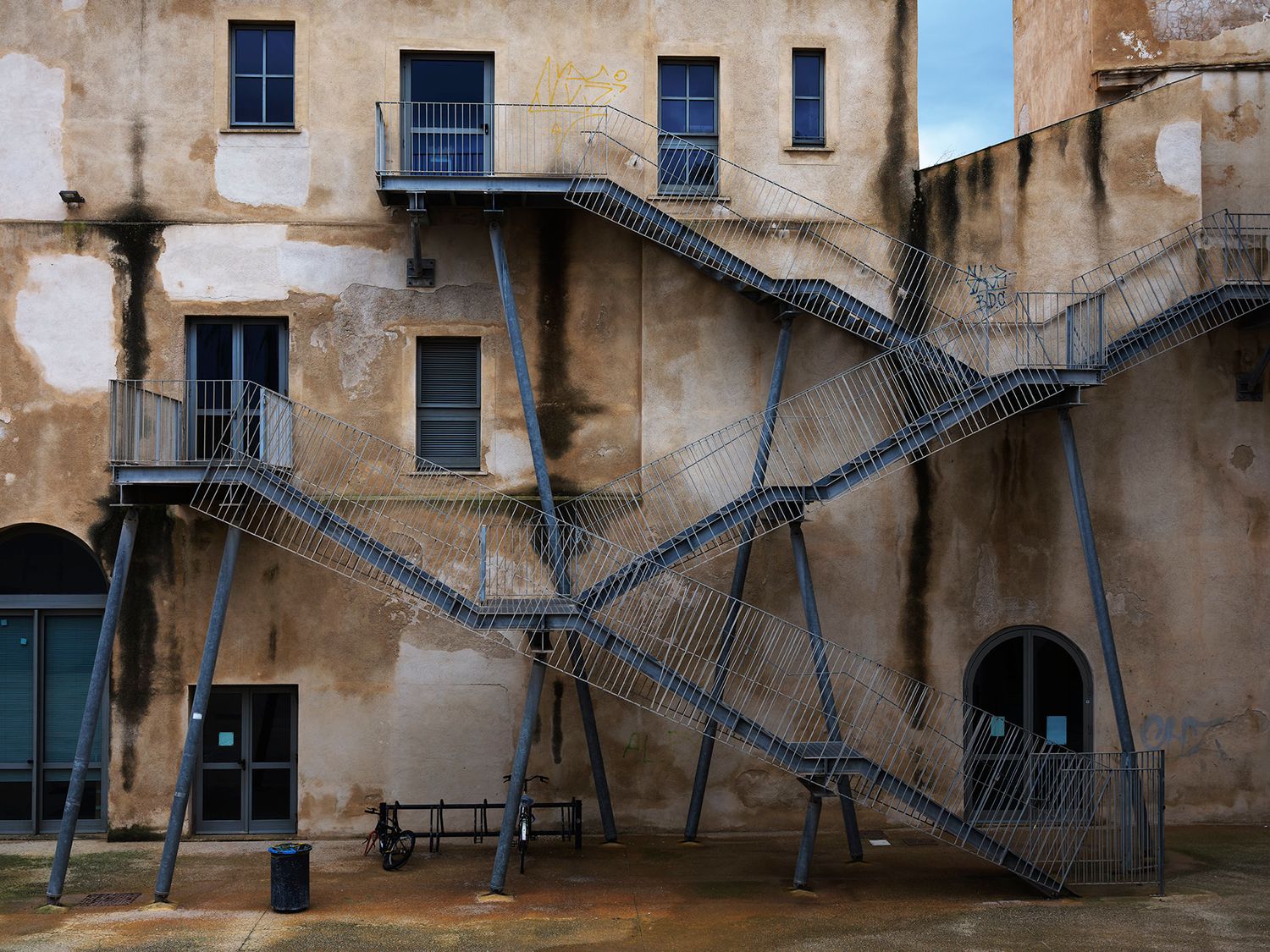 Weathered stone building exterior with zigzag metal staircases and blue-framed windows in an old courtyard.