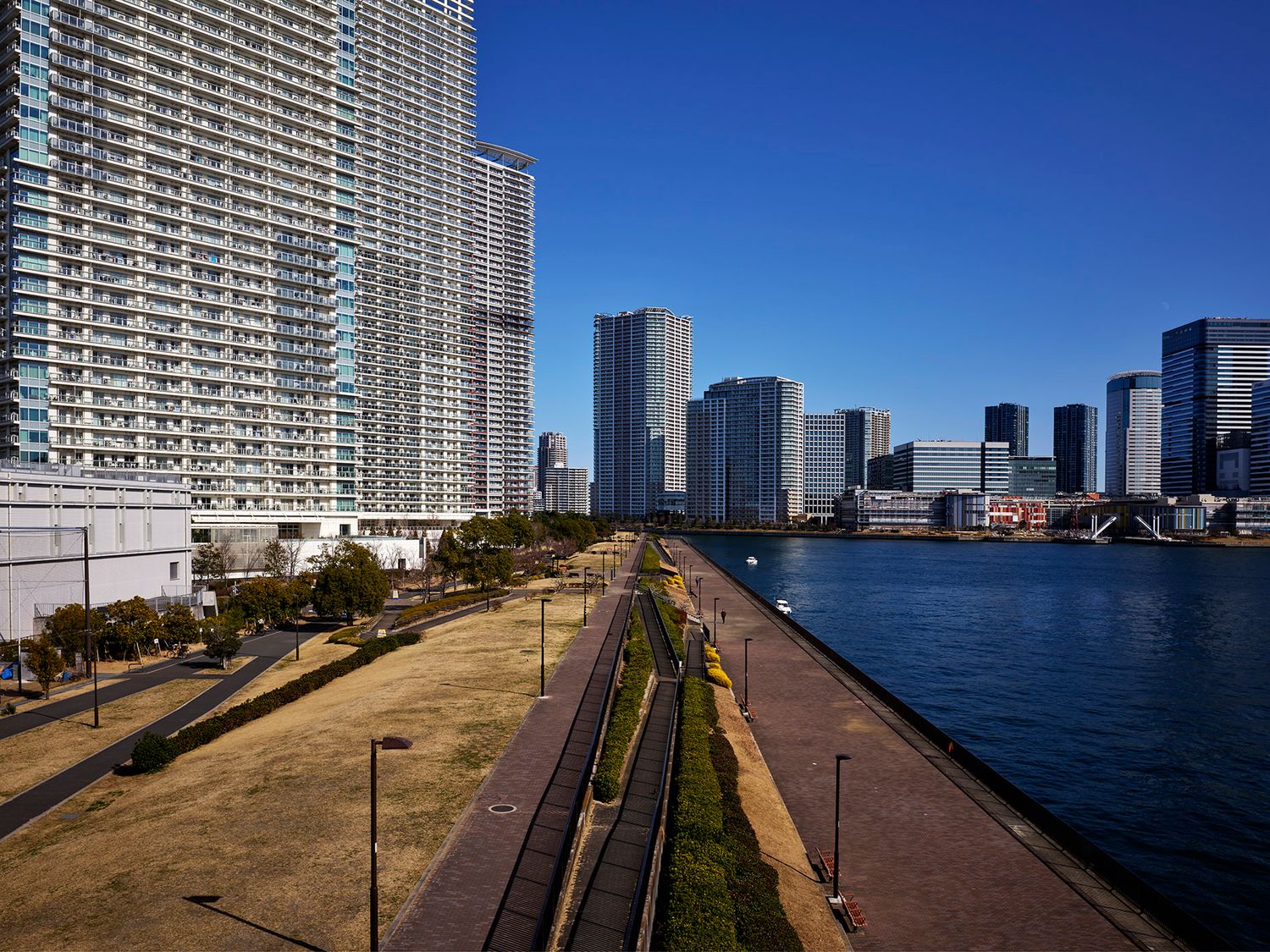 Tokyo waterfront skyline with modern high-rise buildings, riverside promenade, and railway tracks under clear blue sky.