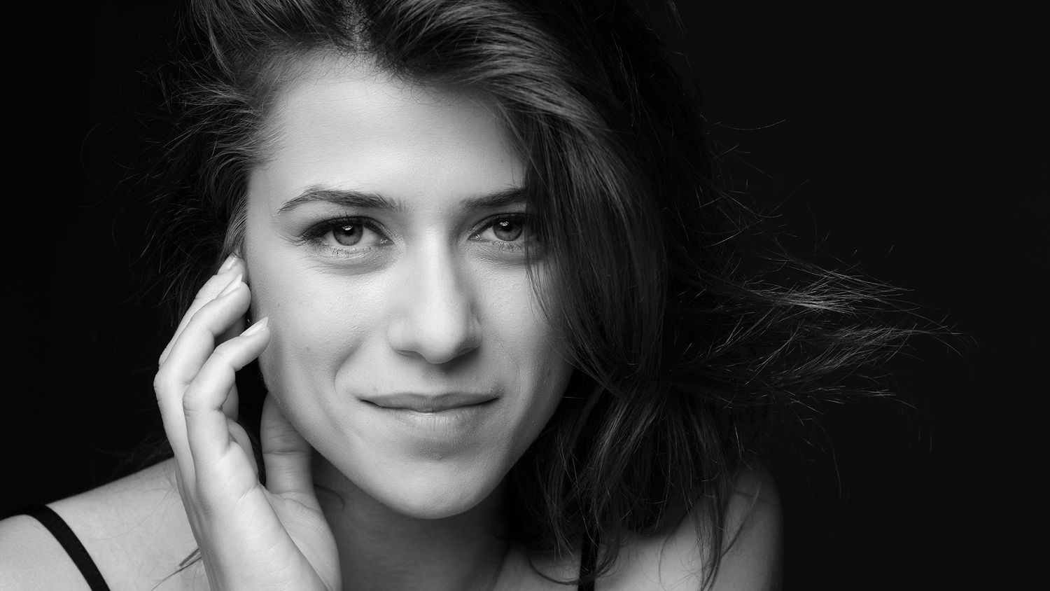 Black and white close-up portrait of a young brunette woman smiling softly touching her cheek.