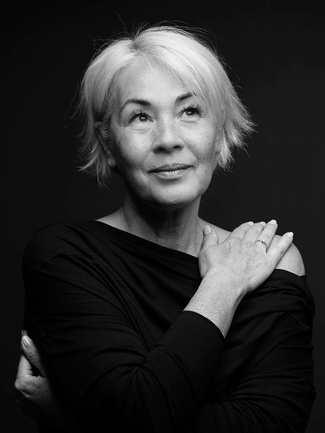 Black and white portrait of an older woman with short blonde hair in a black top, arms crossed.