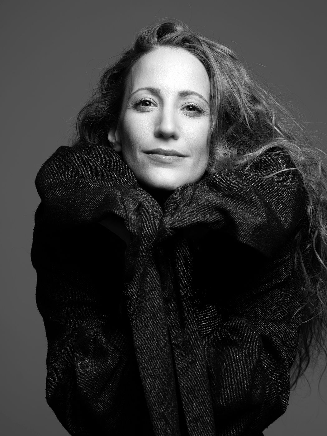 Black and white portrait of a woman with wavy hair wearing an oversized dark coat, smiling softly.