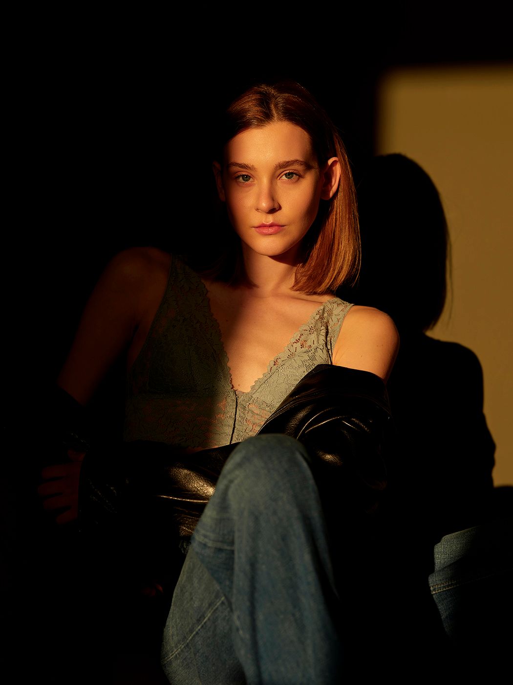 Young woman in lace top and jeans poses dramatically in warm golden light against dark background.
