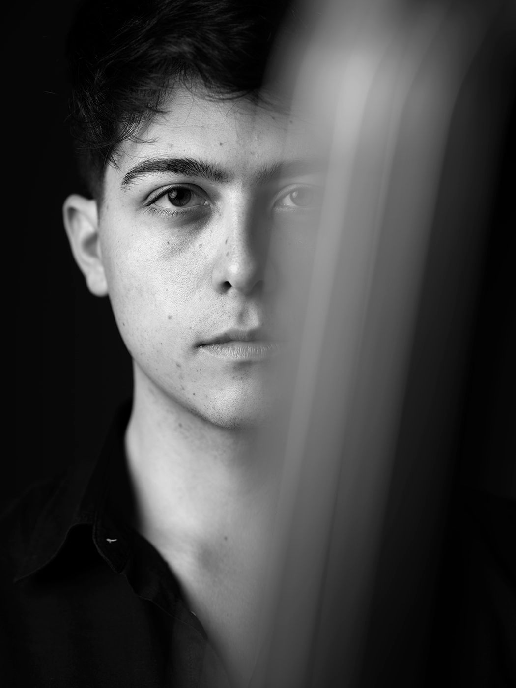 Black and white portrait of a young man with freckles, partially obscured by a blurred foreground element. Mohácsy Lőrinc Ágoston