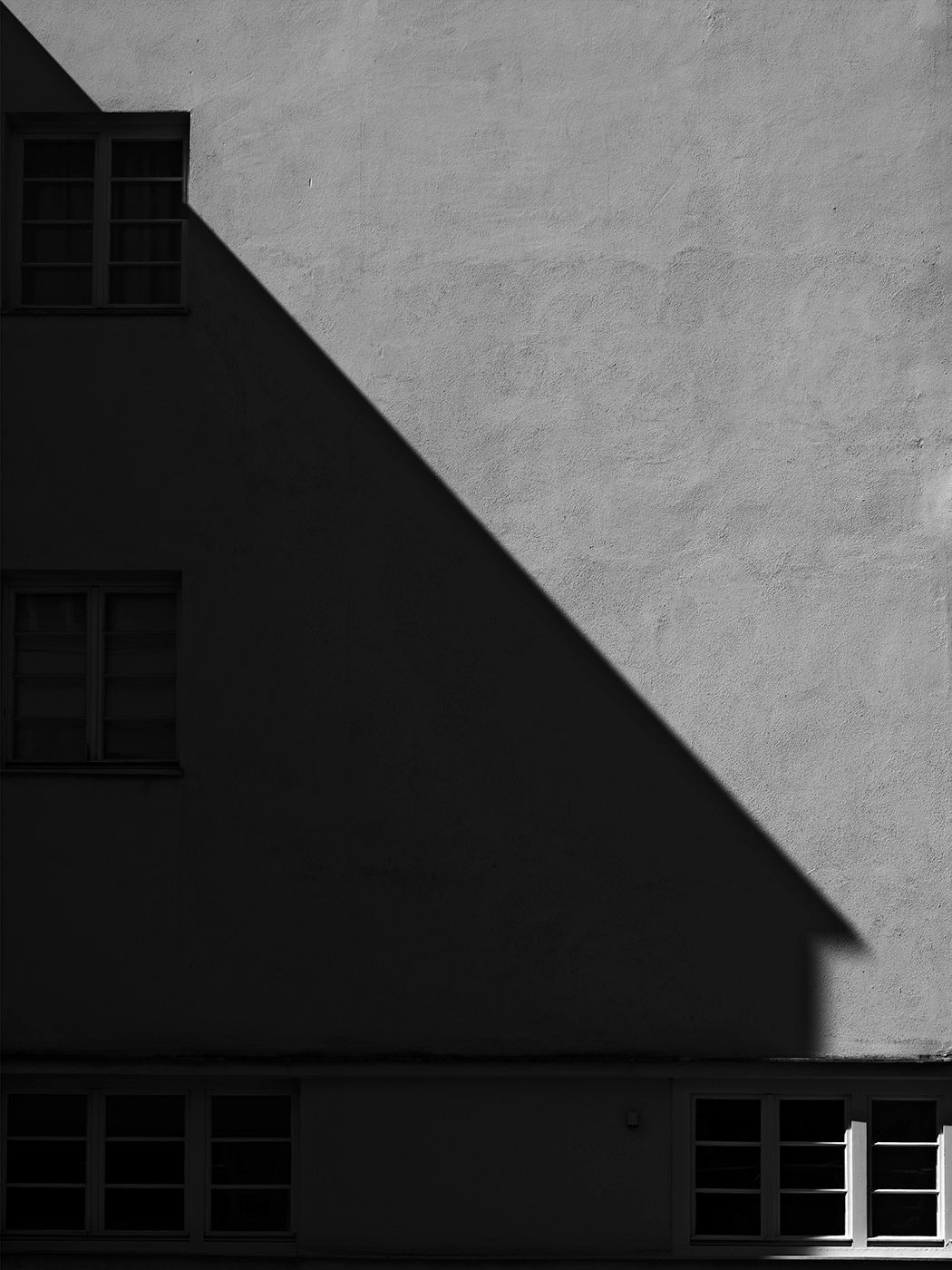 Black and white architectural photo showing a dramatic diagonal shadow cast across a light textured building facade.