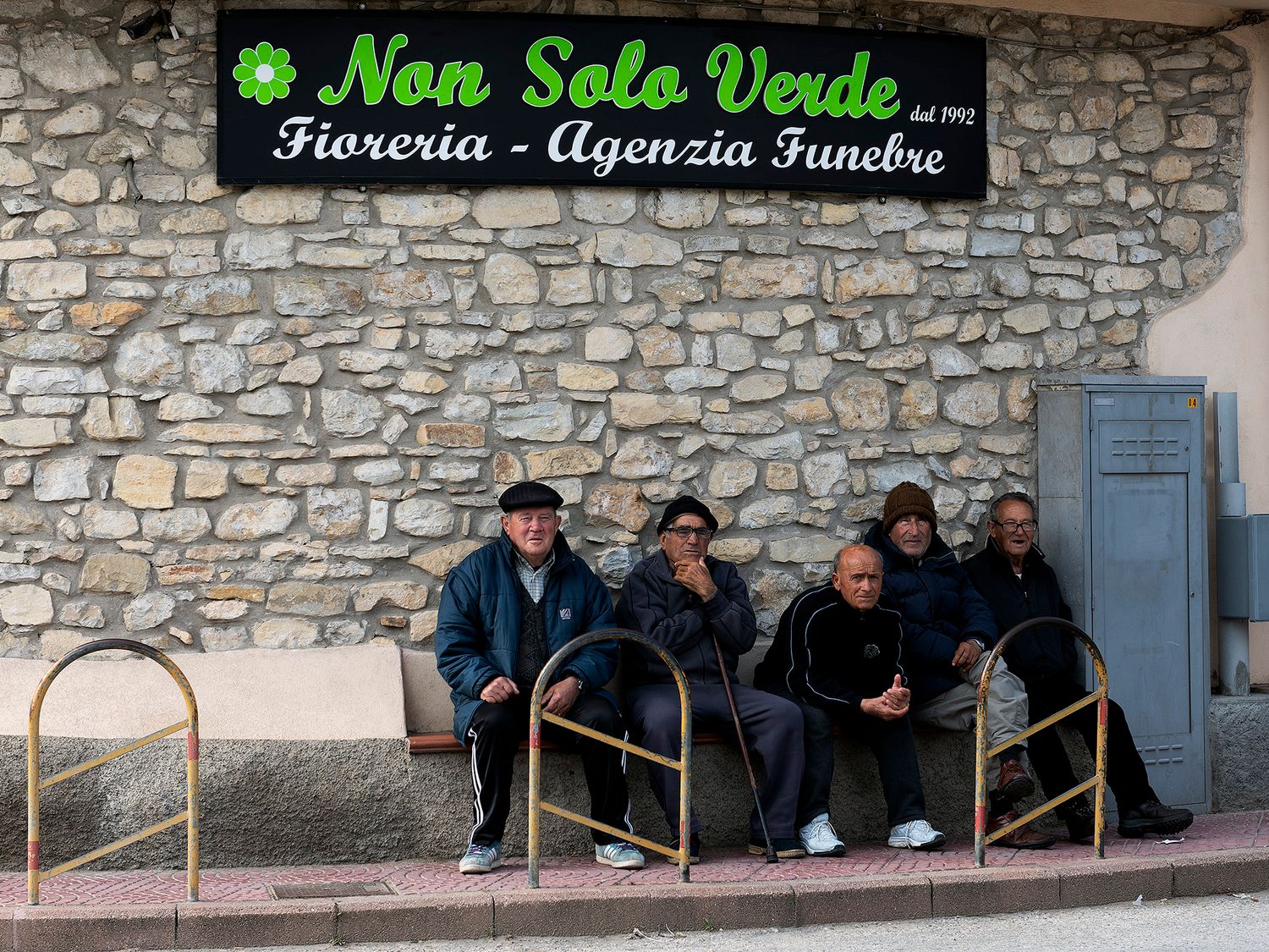 Elderly men sitting on benches outside Non Solo Verde florist and funeral agency in an Italian stone building.