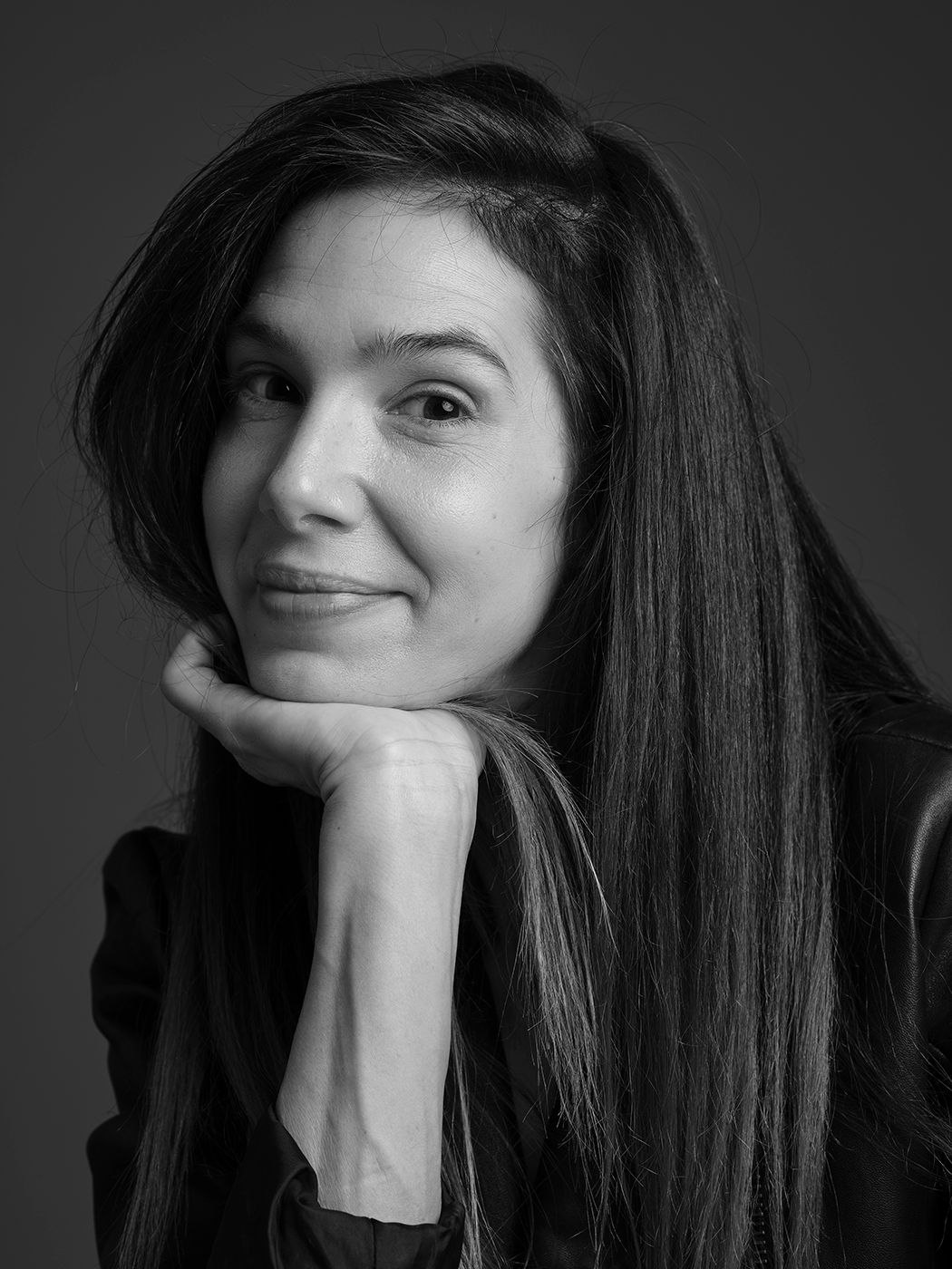 Black and white portrait of a woman with long dark hair, resting chin on hand, smiling softly against gray background.
