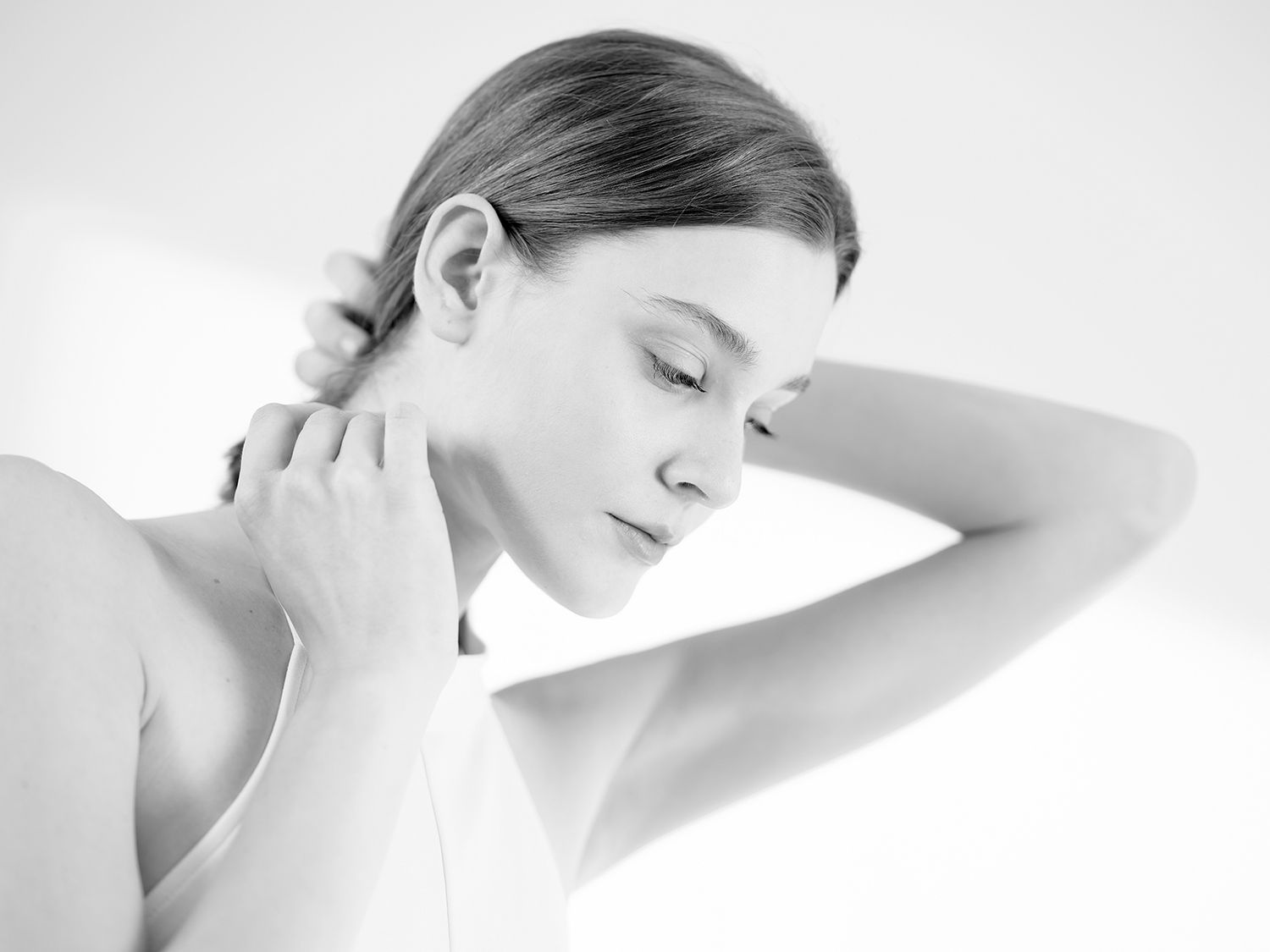 Black and white portrait of a young woman with slicked-back hair, hands raised to her neck, eyes downcast.
