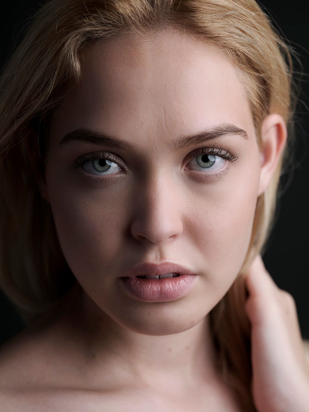 Close-up portrait of a young woman with blue-gray eyes, blonde hair, and natural makeup against a dark background.
