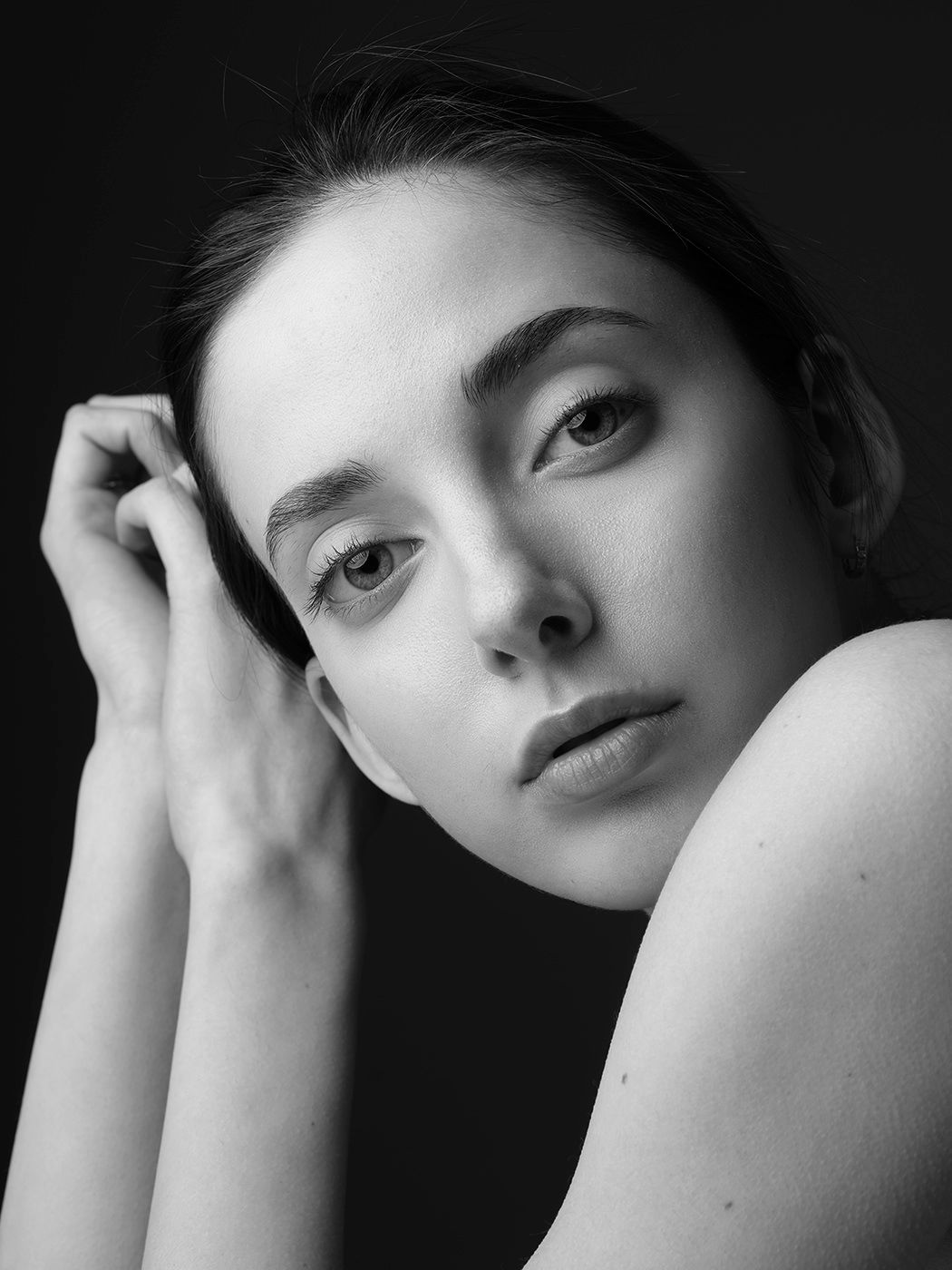 Black and white portrait of a young woman with natural makeup, resting her hand near her face against a dark background.
