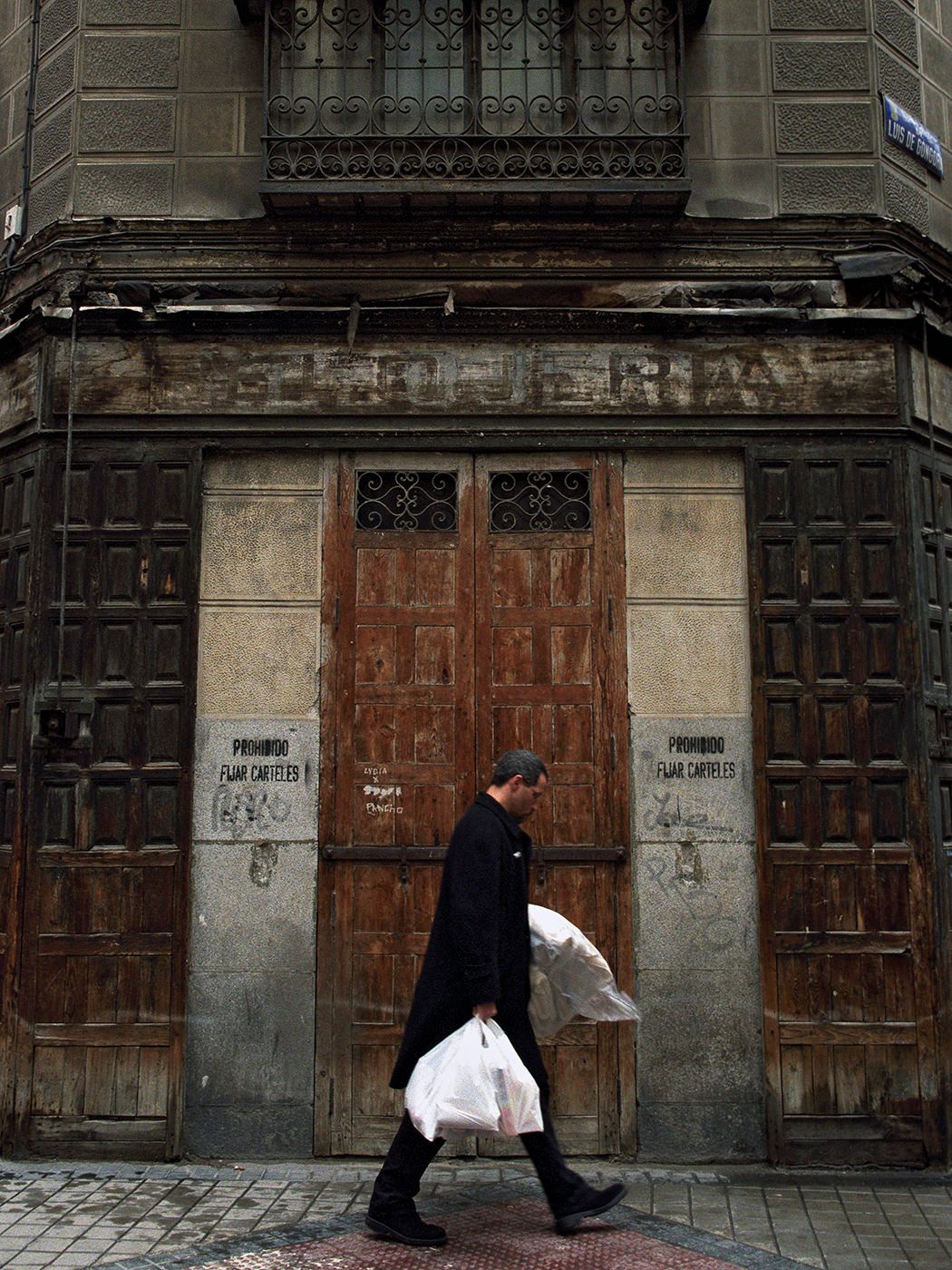 A man in black coat carries shopping bags past a weathered historic building with ornate wooden doors.