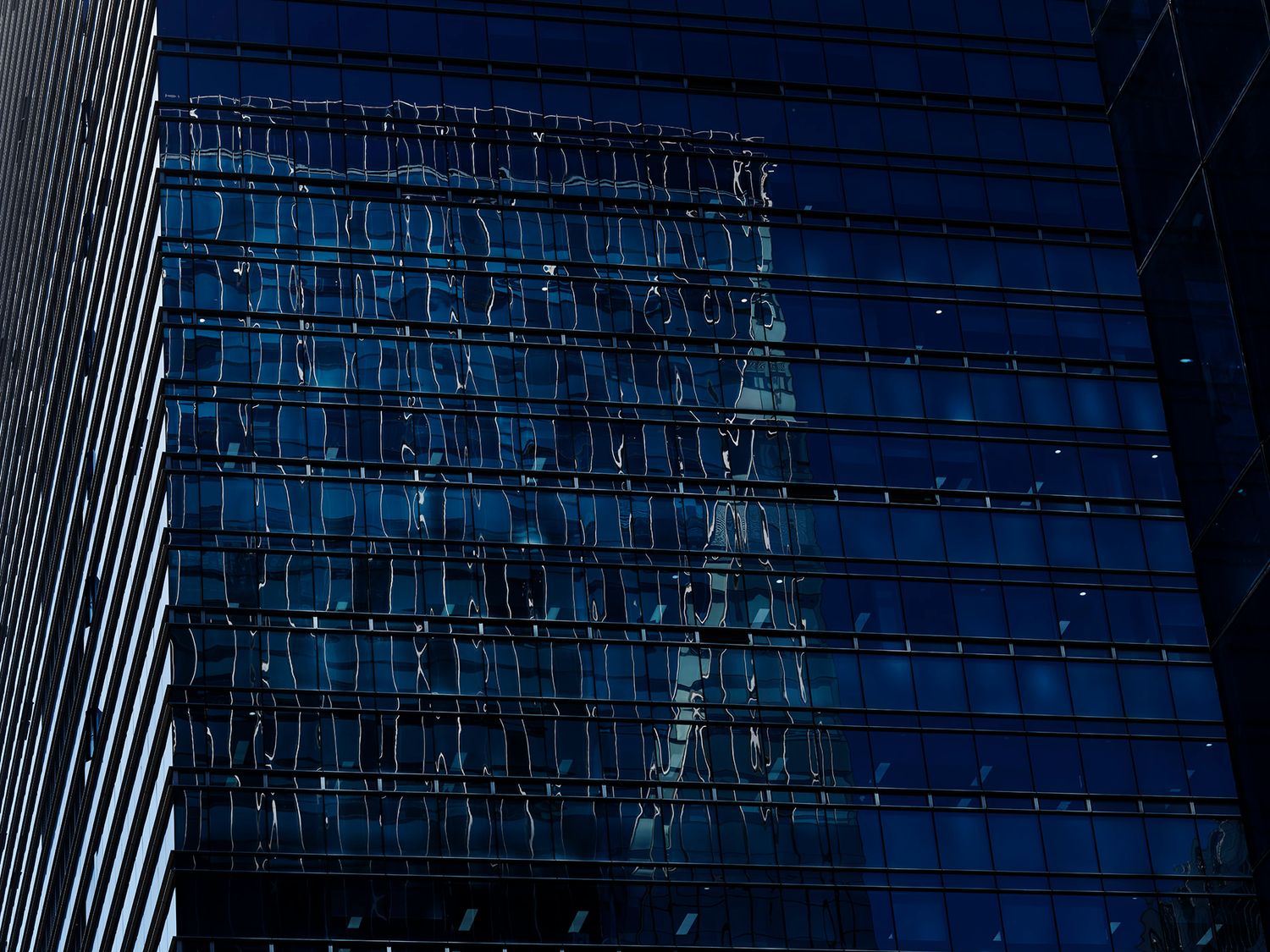 Blue glass skyscraper facade reflecting city buildings at dusk, modern urban architecture with grid pattern windows.