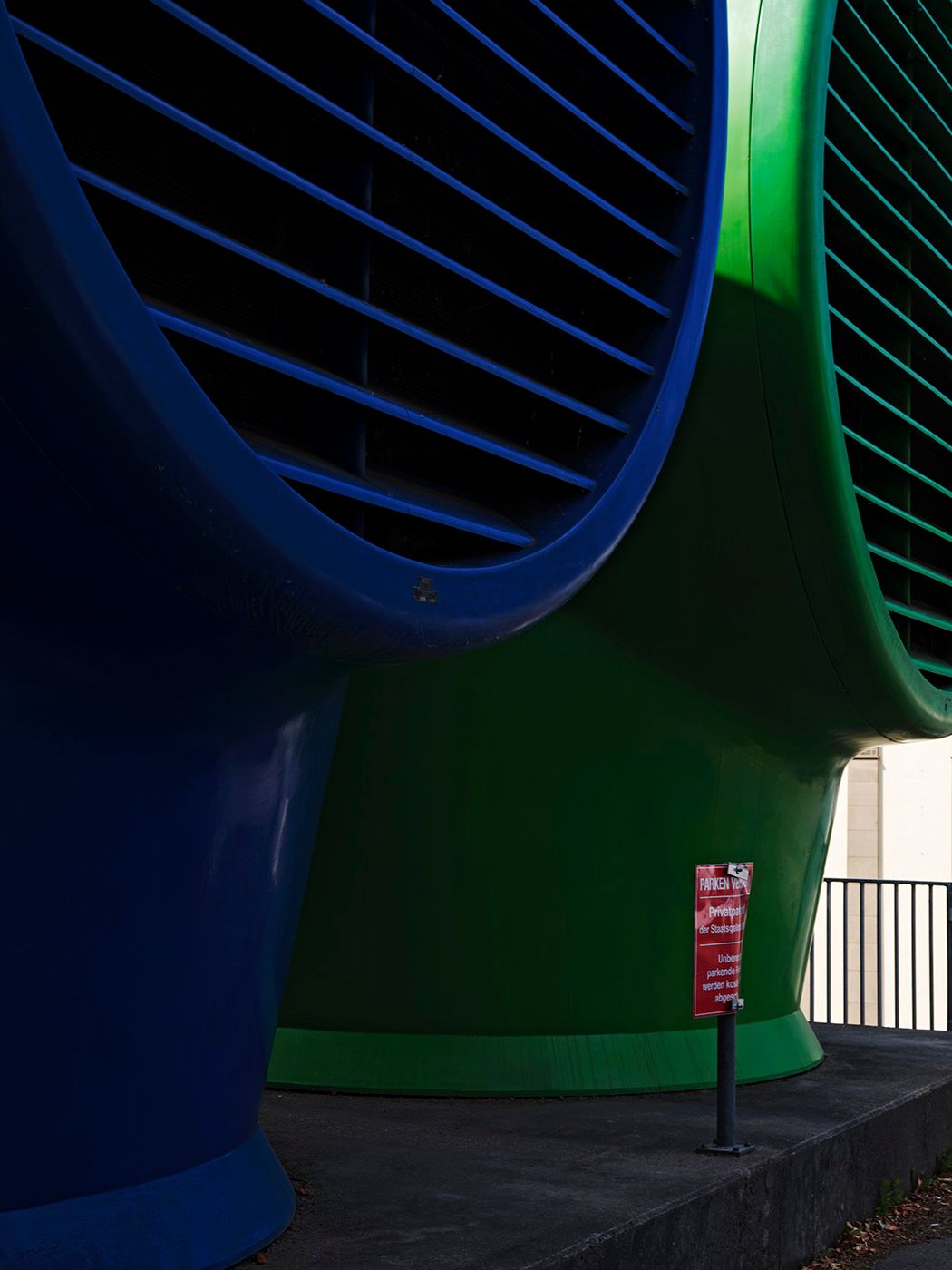 Bold blue and green curved architectural columns with metal grill details beside a red sign and railing.