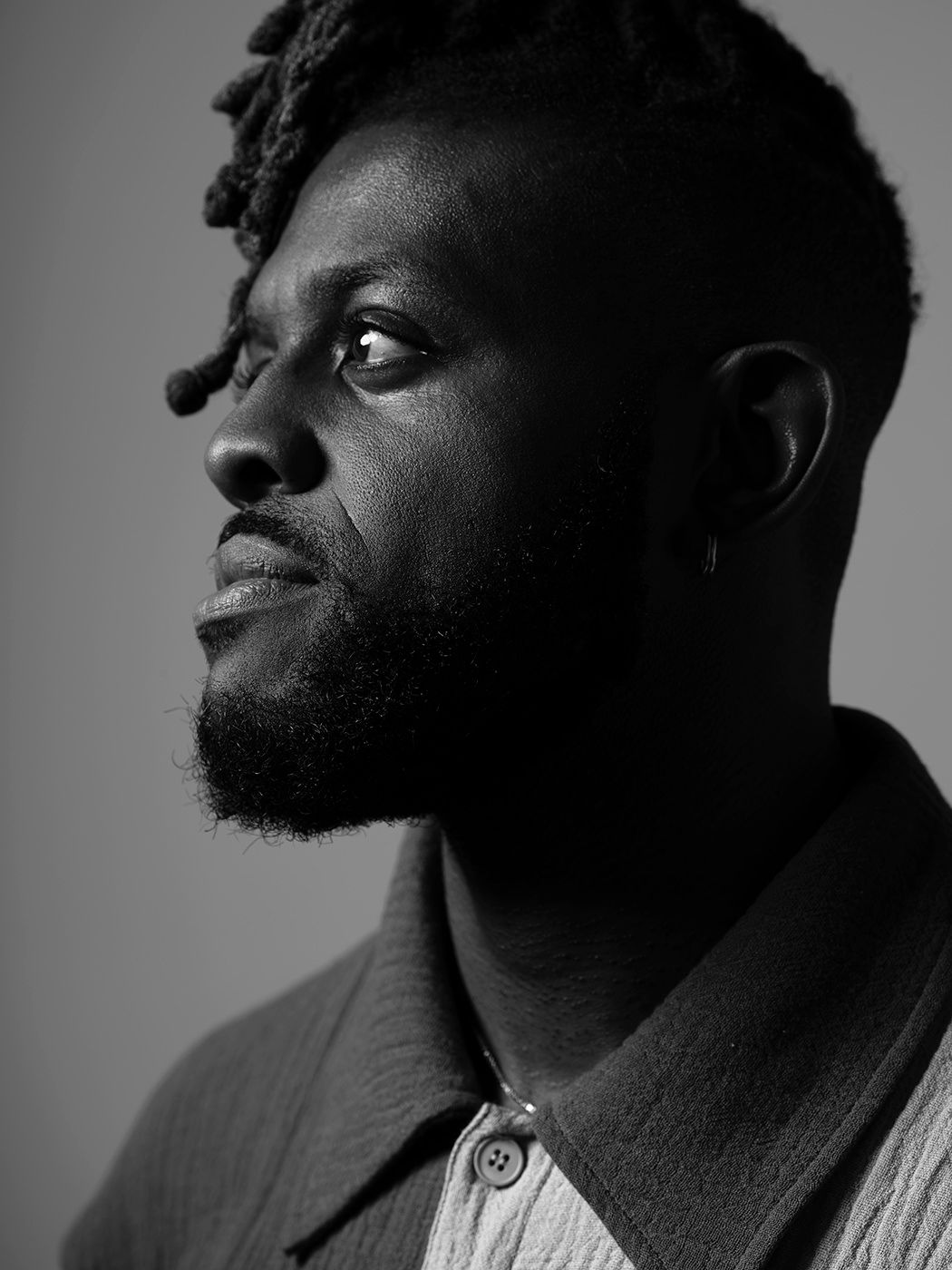Black and white portrait of a young Black man with beard and earring, looking upward in dramatic side lighting. Ashley Henry
