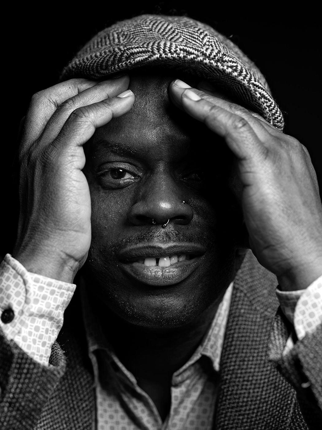 Black and white portrait of a man in a flat cap and blazer, hands framing his face, looking directly at camera. Gregory Hutchinson