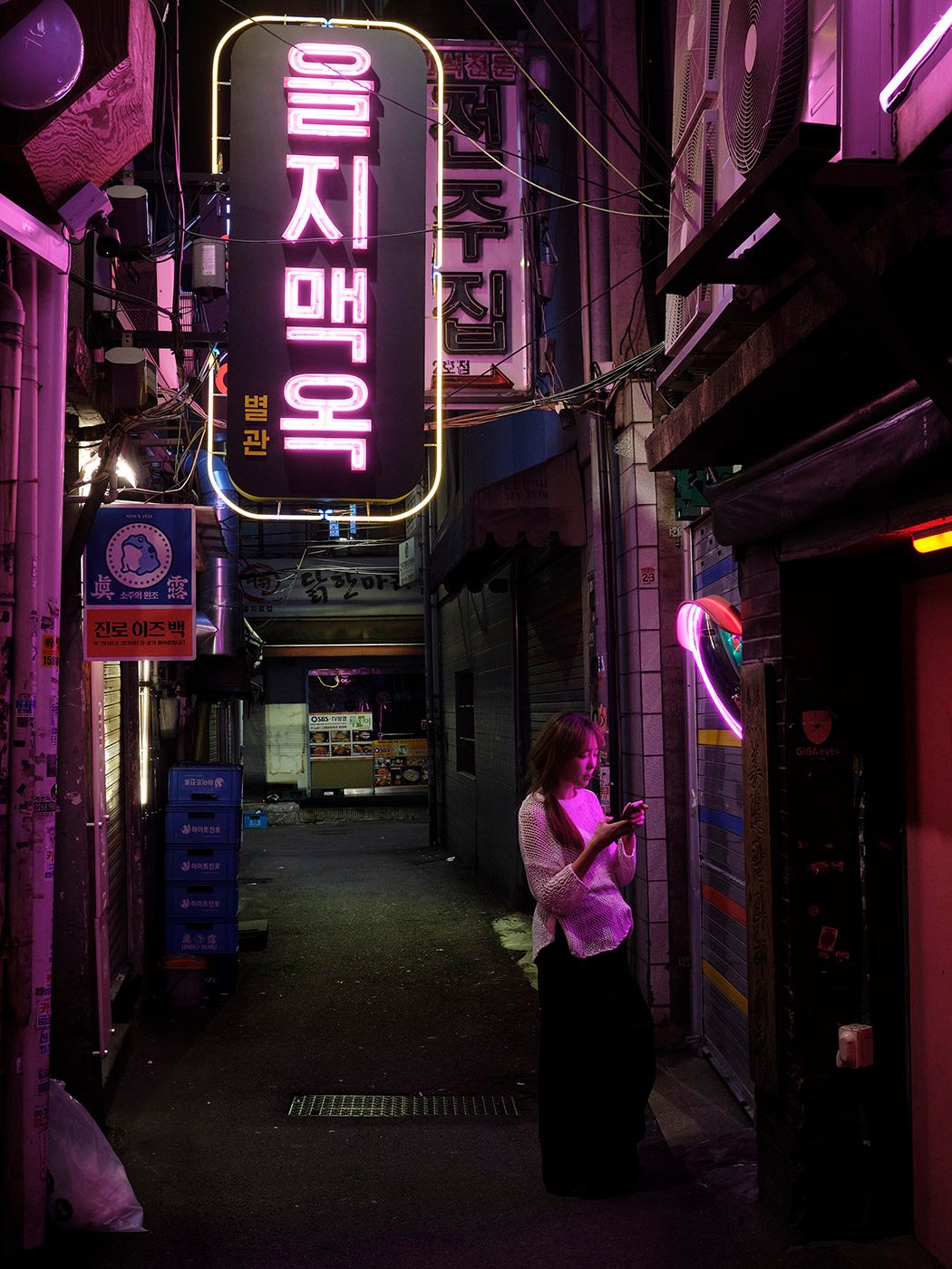 A woman checks her phone in a neon-lit Korean alley at night, surrounded by glowing pink and yellow signs.