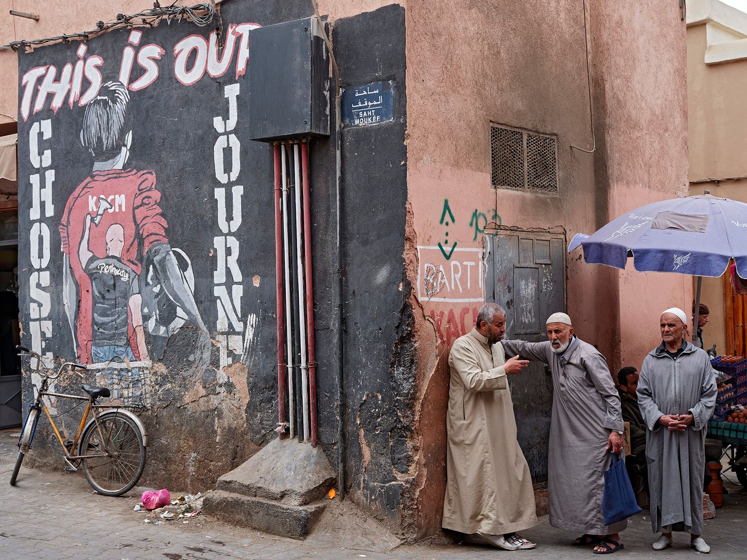 Three men in traditional Moroccan djellabas stand near a colorful street mural in a Marrakech medina alley.