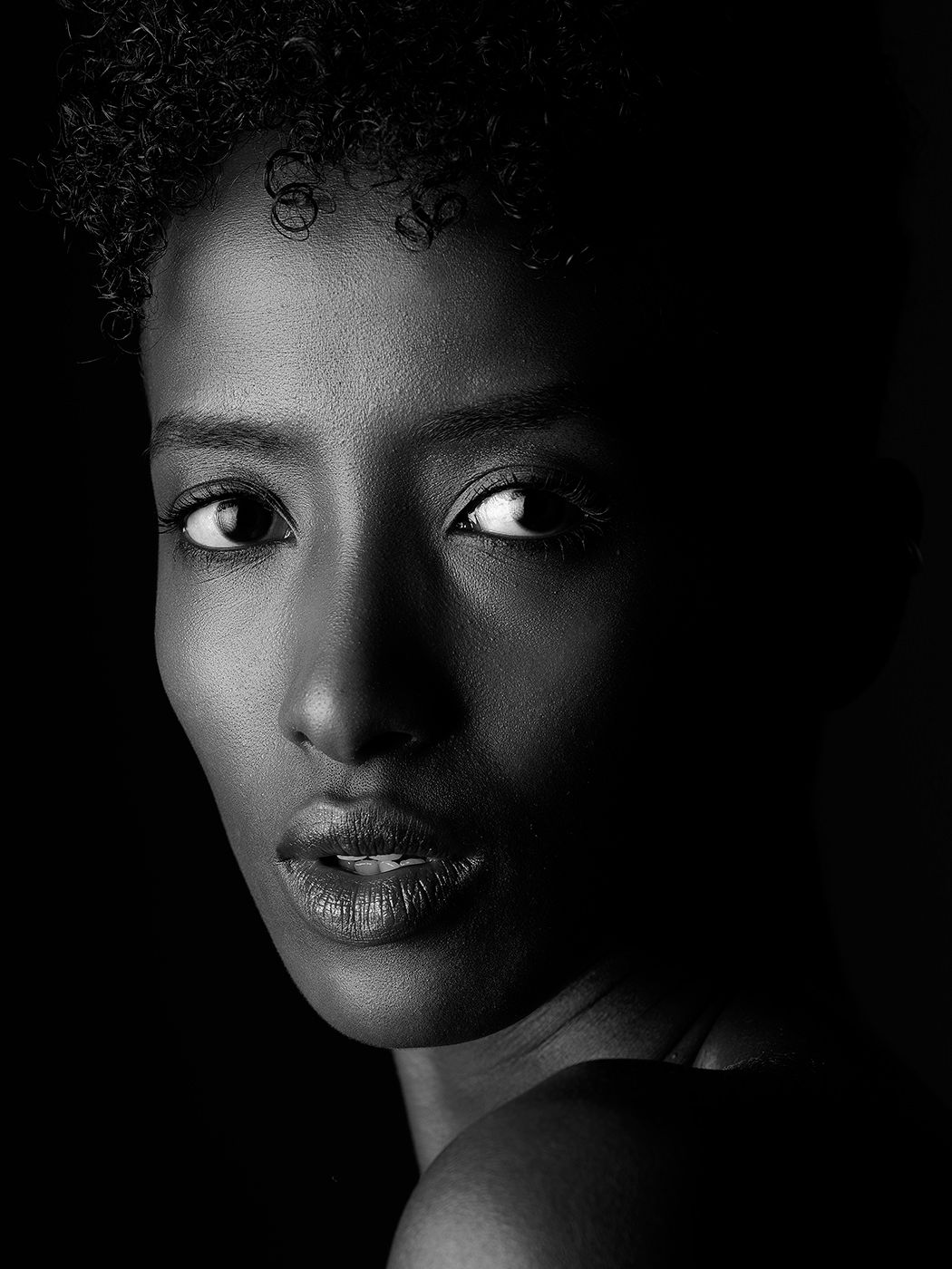 Black and white portrait of a young woman with natural curly hair, striking eyes, and bare shoulders against dark background.