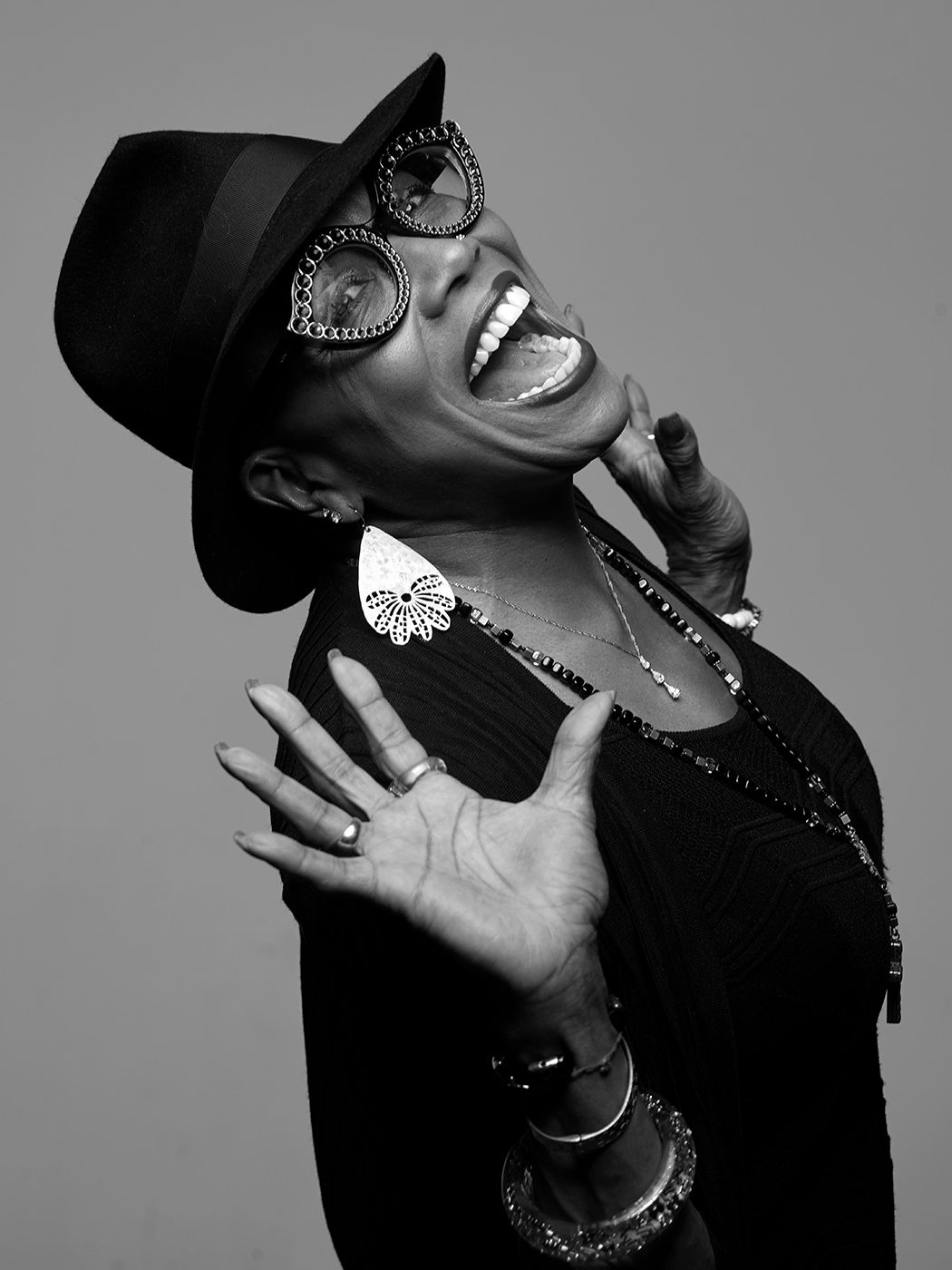 Black and white photo of a joyful woman laughing, wearing a wide-brim hat, large sunglasses, and statement earrings. Dee Dee Bridgewater