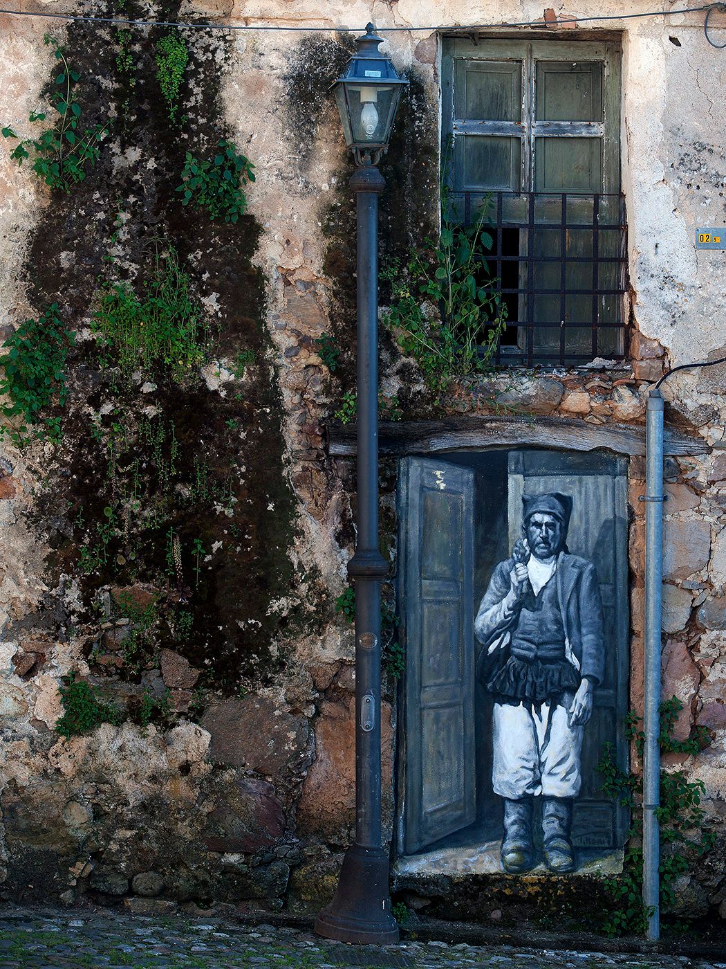 Street art mural of a historical figure painted on an old weathered door beside a vintage lamp post on a rustic stone wall.