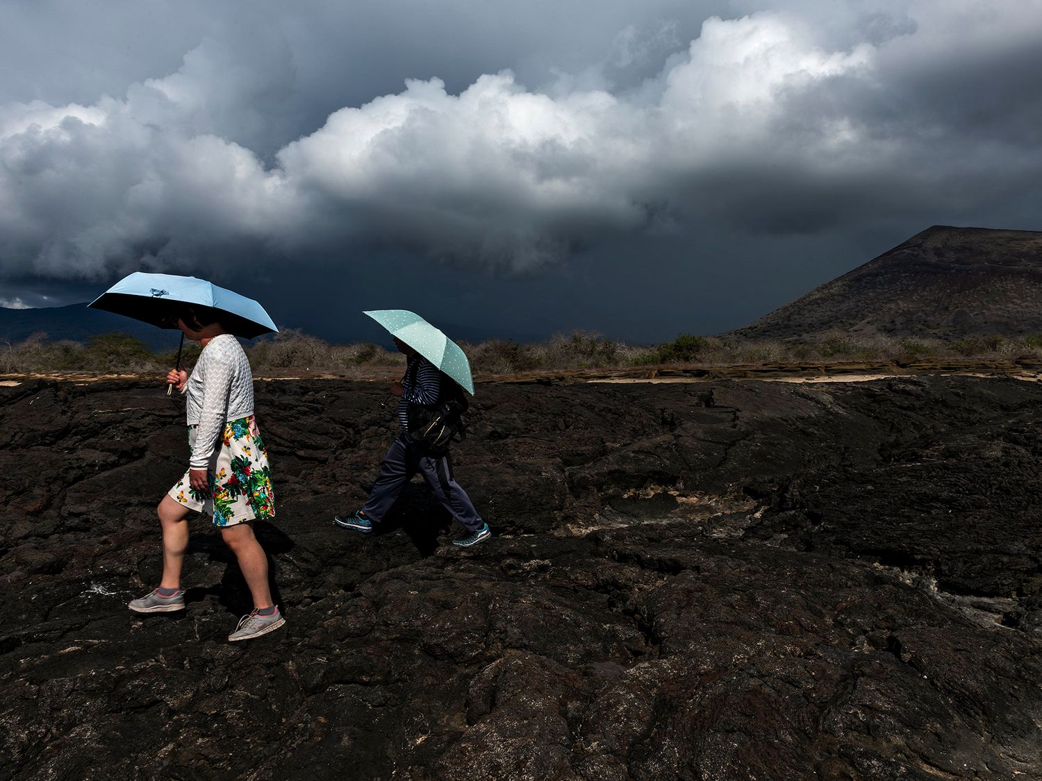 Two people walking with blue umbrellas over dark volcanic rock under stormy clouds.