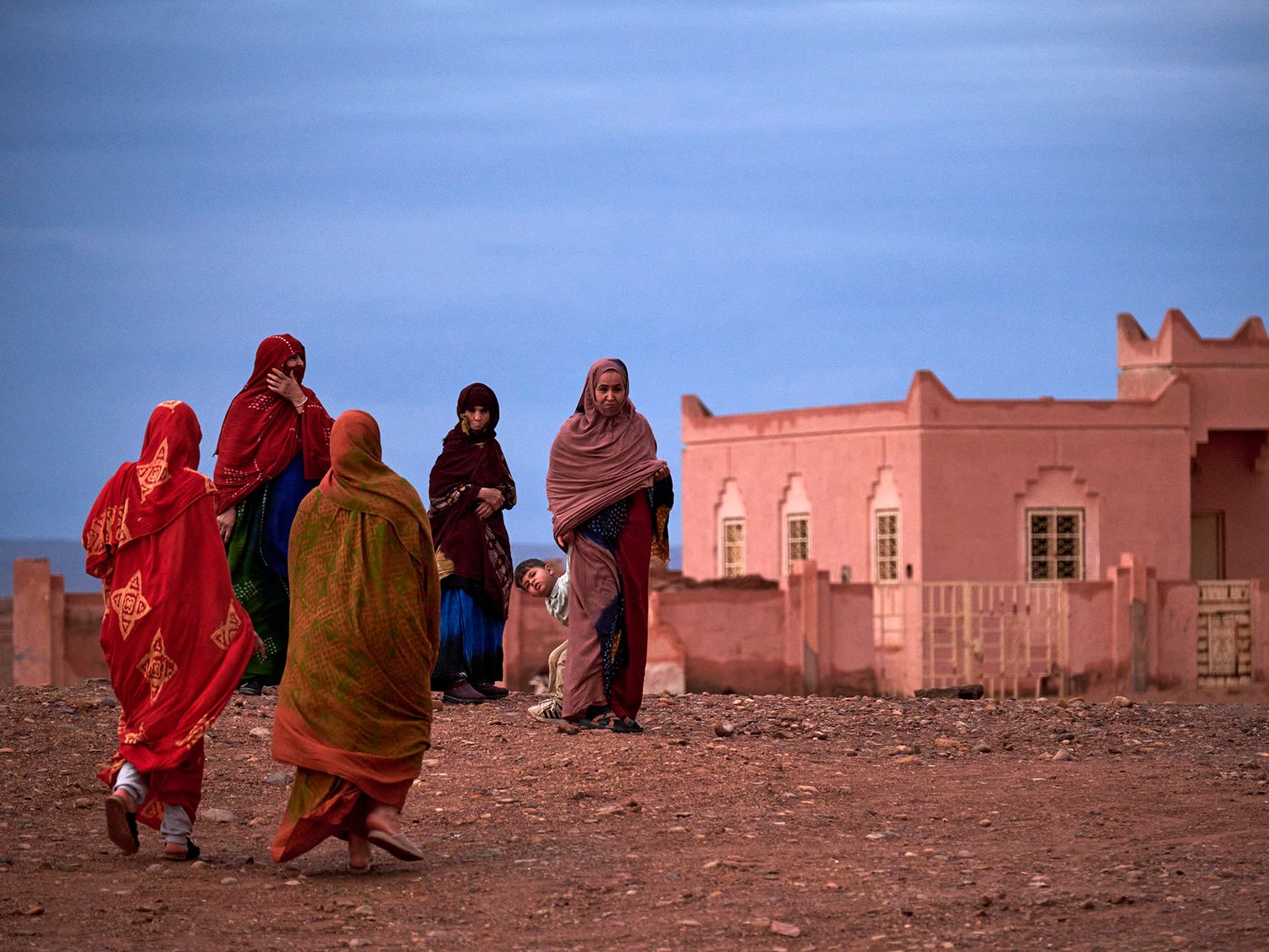 Women in colorful traditional robes walk near a pink adobe building in a North African desert village at dusk.