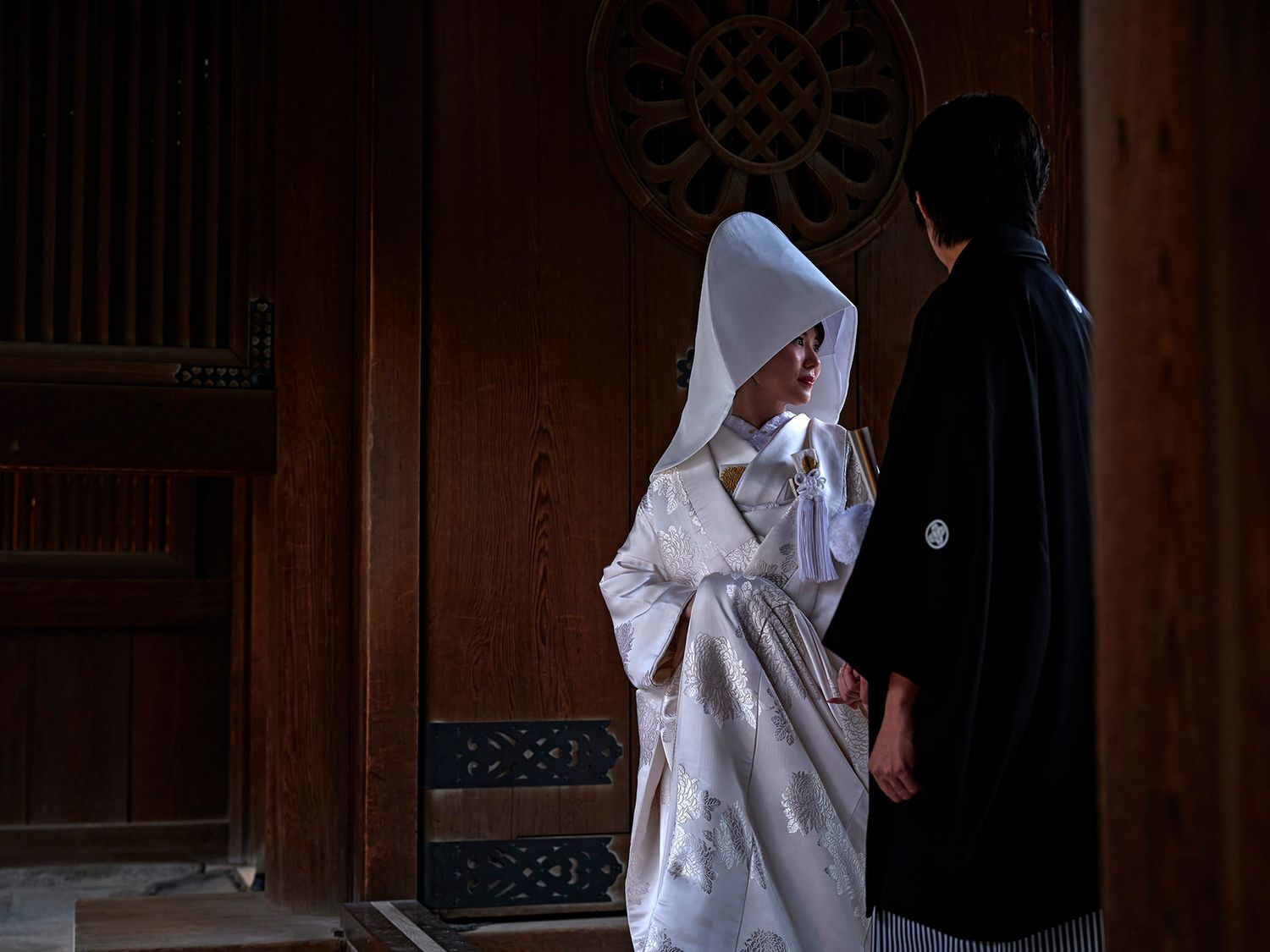 Couple in traditional Japanese wedding attire, bride in white shiromuku kimono and hood, groom in black hakama, in wooden shrine.