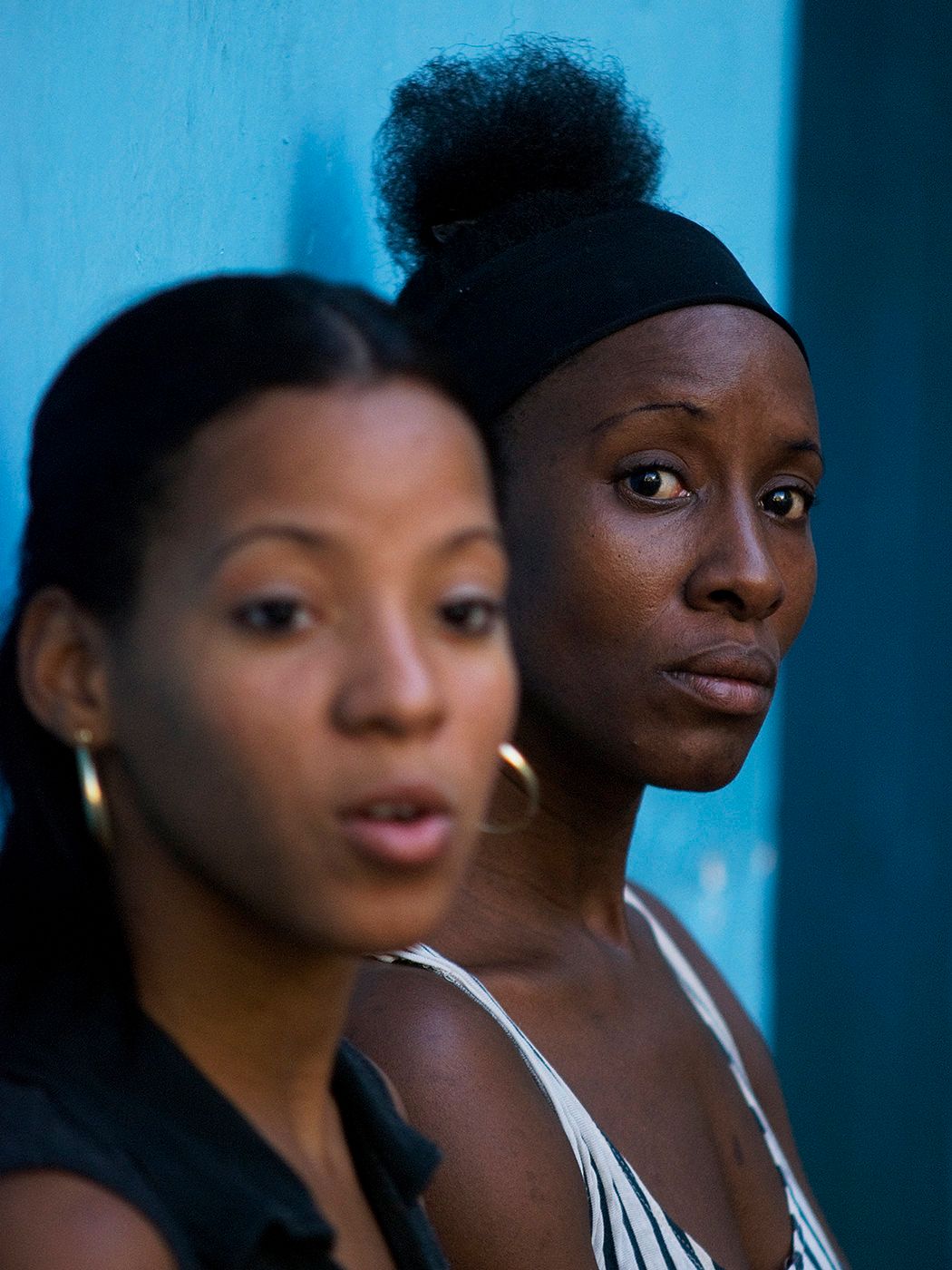Two young Black women posing against a blue wall, one wearing hoop earrings and the other a black headband.