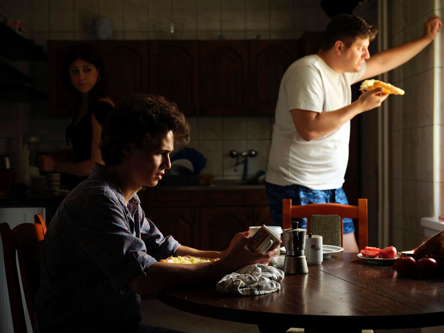 Two young people eating in a dimly lit kitchen with a third person standing nearby.
