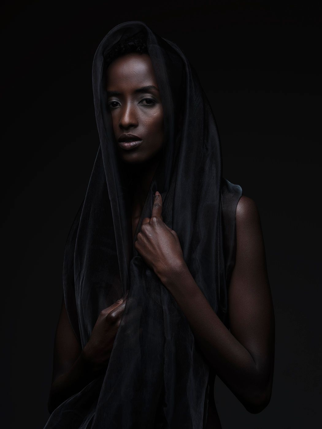 A striking Black woman draped in dark fabric hood poses against a black background in dramatic studio portrait photography.