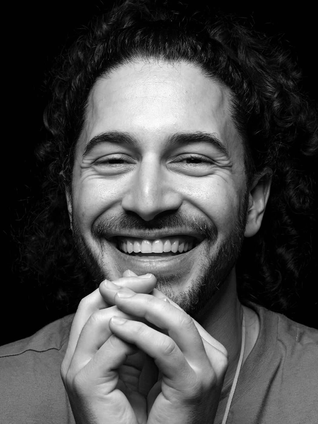 Black and white portrait of a smiling young man with curly hair and beard, hands clasped under chin. Emmet Cohen