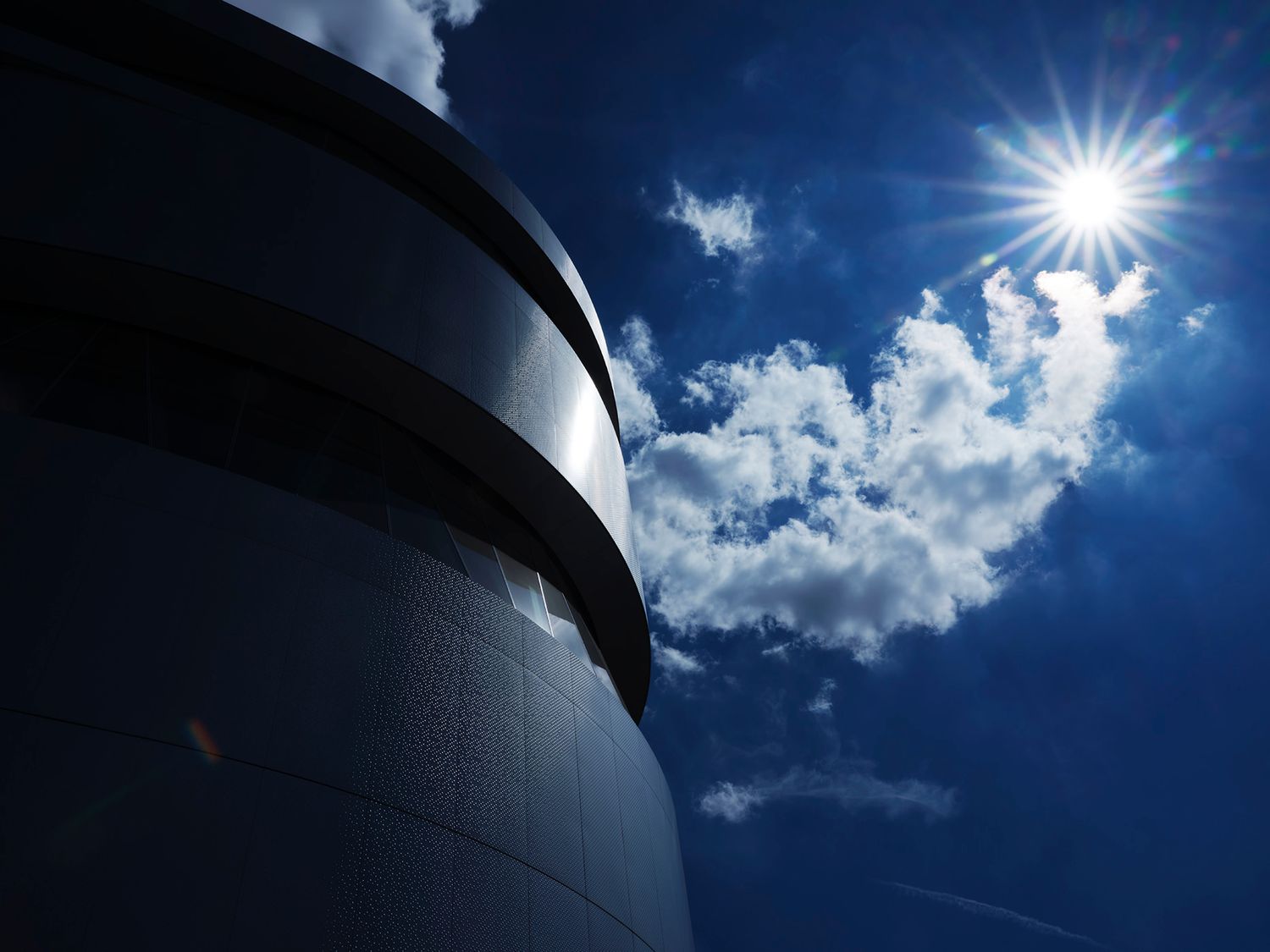 A dramatic low-angle view of a dark cylindrical tower against a vivid blue sky with bright sun and white clouds.