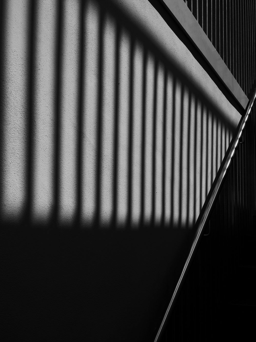 Black and white photo of geometric shadows cast by vertical railings on a textured wall with diagonal lines.