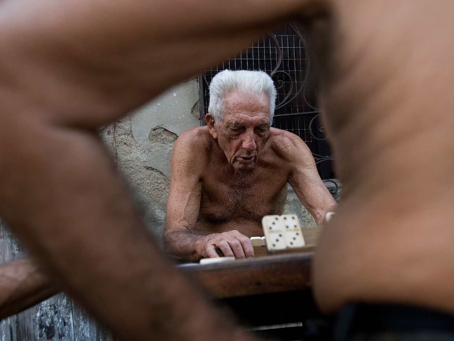 Elderly shirtless man playing dominoes at a table, framed by two people in the foreground.