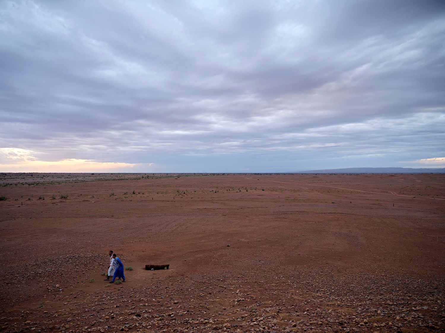 A lone person stands in a vast, arid red desert landscape under a dramatic cloudy sky at dusk.